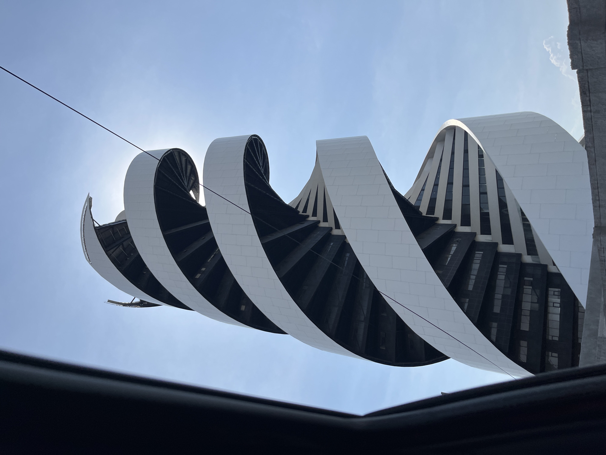 The image shows a low-angle view of an architectural structure in San Bernardino Tlaxcalancingo, Mexico, as perceived from beneath a sunroof. The building features a distinctive design, with spiraling white and black elements that curve upwards towards the sky. The white sections appear to be clad in rectangular tiles. The design reveals glimpses of windows behind the spiraling facade. The sky is a clear blue, with the sun partially obscured by one of the architectural elements, creating a lens flare effect. A thin, dark wire or cable is visible against the sky. The frame of the sunroof occupies the bottom of the image, suggesting the perspective of someone looking up from inside a vehicle. A concrete or stone structure is visible on the right side of the frame.