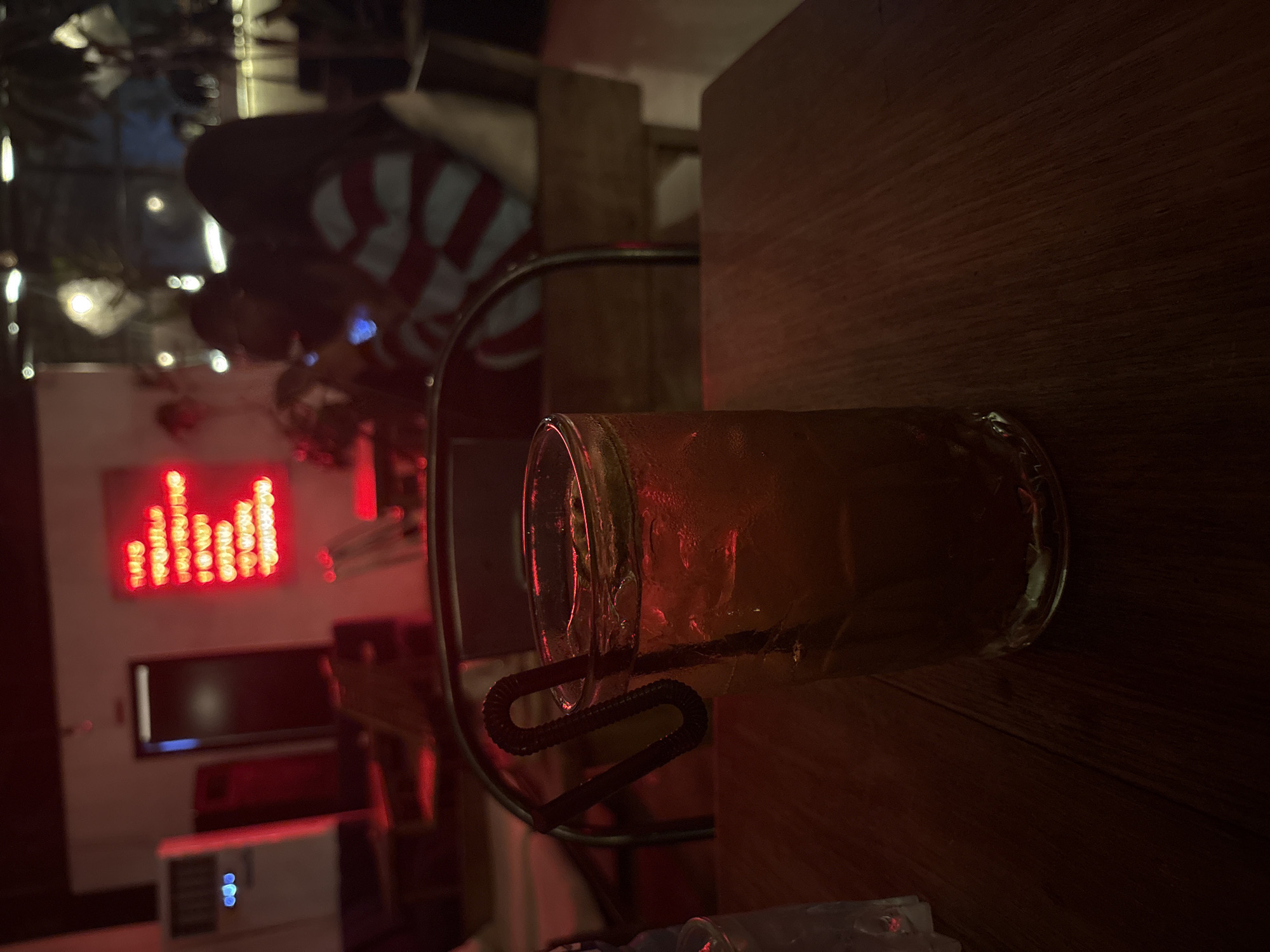 A slightly tilted, medium-angle shot depicts a bar scene, likely in Abuja, Nigeria, given the context. In the foreground, on a dark wooden surface, sits a clear glass filled with a light-colored beverage, complete with a dark, curly straw. Condensation is visible on the glass. Behind the glass is a metal chair frame. In the blurred background, illuminated by ambient light, are figures and a wall with a red neon sign. A dark doorway is also visible. The overall lighting is dim and warm, creating an intimate ambiance.
