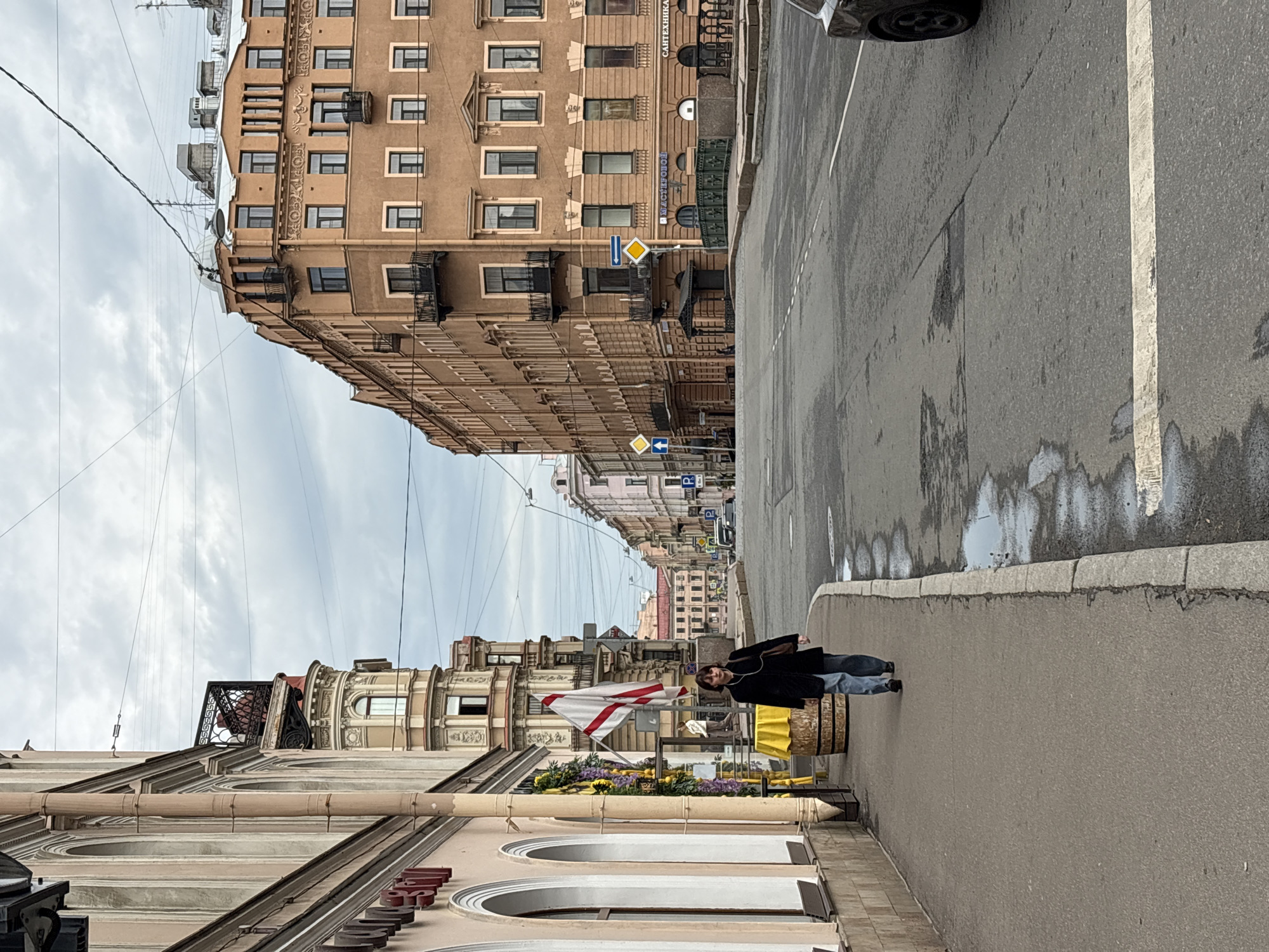 A street scene in Saint Petersburg, Russia, shows buildings lining a street on either side, receding into the distance. A pedestrian, dressed in dark clothing and light-colored jeans, stands on the sidewalk next to a display with flowers and a white flag with a red stripe. A utility pole with numerous wires stretches across the sky above the buildings. The street is wet, reflecting light. A brown building with multiple windows is on the right side. A lighter-colored building with arched windows is on the left.