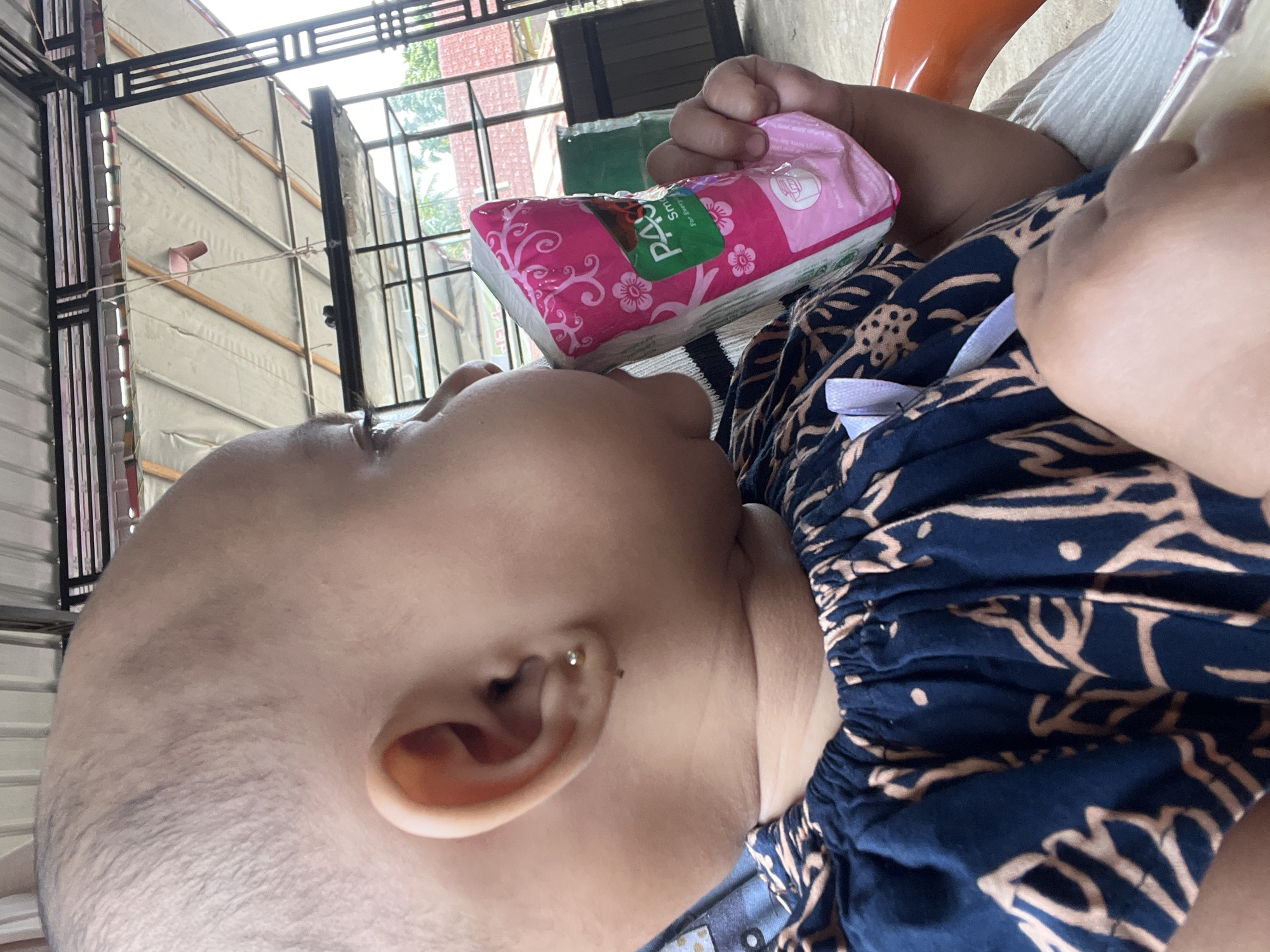A close-up, profile view shows a young child, likely an infant or toddler, with a shaved head and dark brown eyes. The child is wearing a dark blue garment with a light brown pattern and a white ribbon at the neckline. The child is holding a pink and white packaged item, possibly a juice box or pouch, and appears to be drinking from it. The child has a small stud earring in their left ear. The background suggests an outdoor or semi-outdoor setting, possibly a patio or covered area, with a corrugated metal roof overhead supported by black metal beams. Some greenery and buildings are visible in the distance. The scene is well-lit, suggesting daytime. The environment appears to be a residential area in Seumirah, Indonesia.
