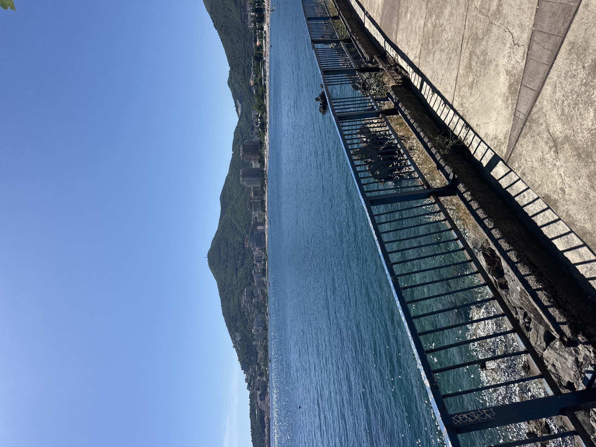 View of the Adriatic Sea from a coastal promenade in Budva, Montenegro. The sea extends to the left of the frame, bounded by the coastline and mountains on the left. The near right of the frame shows a portion of the promenade, including a railing, curb, and sidewalk. The sky is clear and blue. The water glistens with sunlight. Buildings are visible along the coastline on the mountain. The railing casts shadows onto the curb and sidewalk.