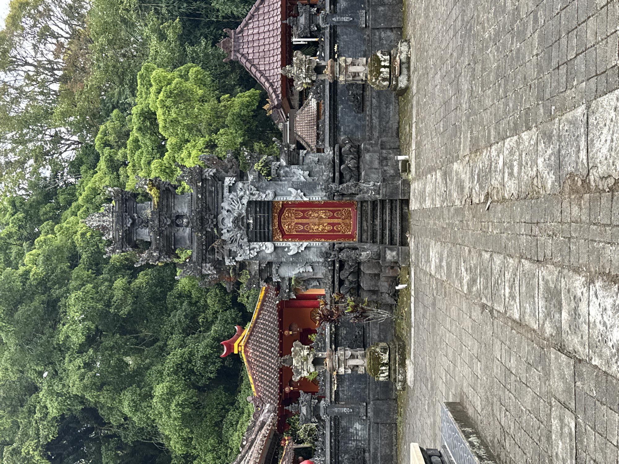 The image presents a serene view of a traditional Balinese temple complex set against a backdrop of lush tropical greenery. The central feature is an intricately carved dark grey stone gateway, likely a Candi Bentar, adorned with elaborate reliefs and statuary. A prominent, ornate red and gold double door, featuring delicate golden patterns, is visible at the top of a short set of steps within this gateway.
Flanking the main gateway are smaller temple pavilions with distinctive reddish-brown tiled roofs, supported by dark stone bases and further embellished with carvings. Stone pedestals or altars, some lightly covered in moss, are positioned in front of these structures and along the paved pathway.
The background is filled with a dense, vibrant green forest of tall trees, emphasizing the natural and secluded setting. The lighting is soft and diffused, suggesting an overcast day. A wide, light grey paved courtyard or pathway made of rectangular stone blocks occupies the foreground and extends towards the right side of the frame, leading up to the temple entrance.
No people or discernible human activity are present, lending a tranquil atmosphere to the scene. The architectural style, materials, and vegetation are characteristic of cultural sites in Bali, consistent with the location context of Baturiti, Indonesia. There is no visible text in the image.