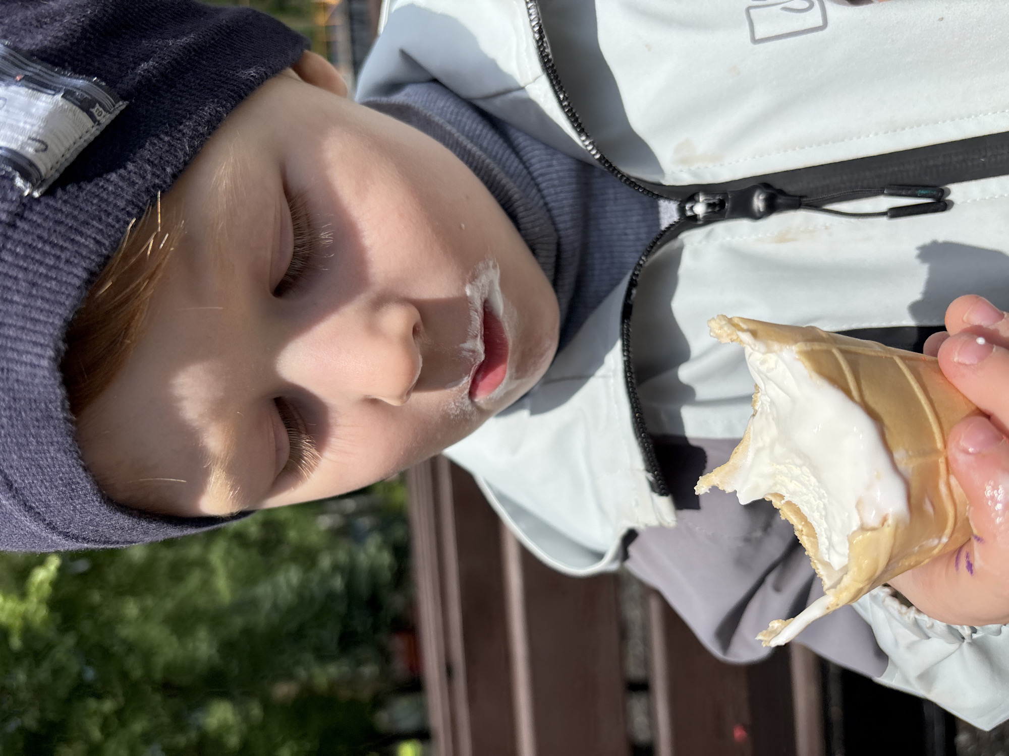 A young child is seen close-up, angled to the left, holding and eating an ice cream cone. The child wears a dark blue knitted hat and a light grey jacket with a visible zipper. There is melted ice cream around the child's mouth. The background shows a blurred scene of green trees and a brown wooden structure. While the location is not explicitly mentioned in the image itself, it can be inferred that the child is outdoors, potentially in a park or recreational area. Since the location is stated as Izhevsk, Russia, that the image may have been taken in Izhevsk, Russia.