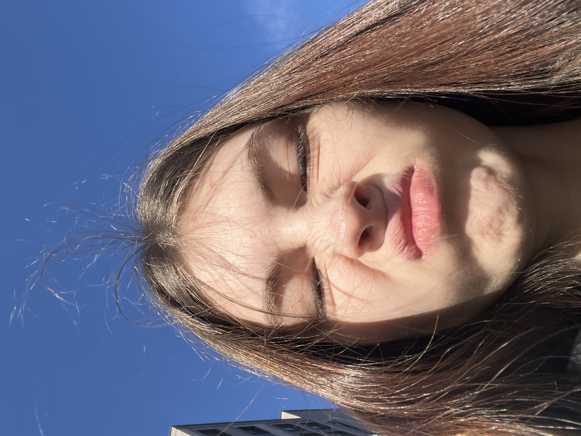 A close-up, low-angle portrait features a young woman with long, dark brown hair against a bright, clear blue sky. Her head is tilted, and her face is positioned to look upwards towards the camera or the sun. She has her eyes tightly squinted, her nose is visibly wrinkled, and her lips are slightly puckered, suggesting either a playful expression or a strong reaction to the intense sunlight.
Her hair, illuminated by the sun, shows off various reddish-brown highlights and fine, flyaway strands, particularly on the left side of the frame. Direct sunlight casts strong shadows across her face, highlighting the contours of her nose and cheeks. The background is almost entirely a vibrant, unclouded blue sky, indicating sunny weather during the day. In the bottom left corner, a portion of a modern building with dark windows is faintly visible, suggesting an urban environment. The image conveys a candid and lighthearted moment, set outdoors in Saint Petersburg, Russia. No text is visible in the image.