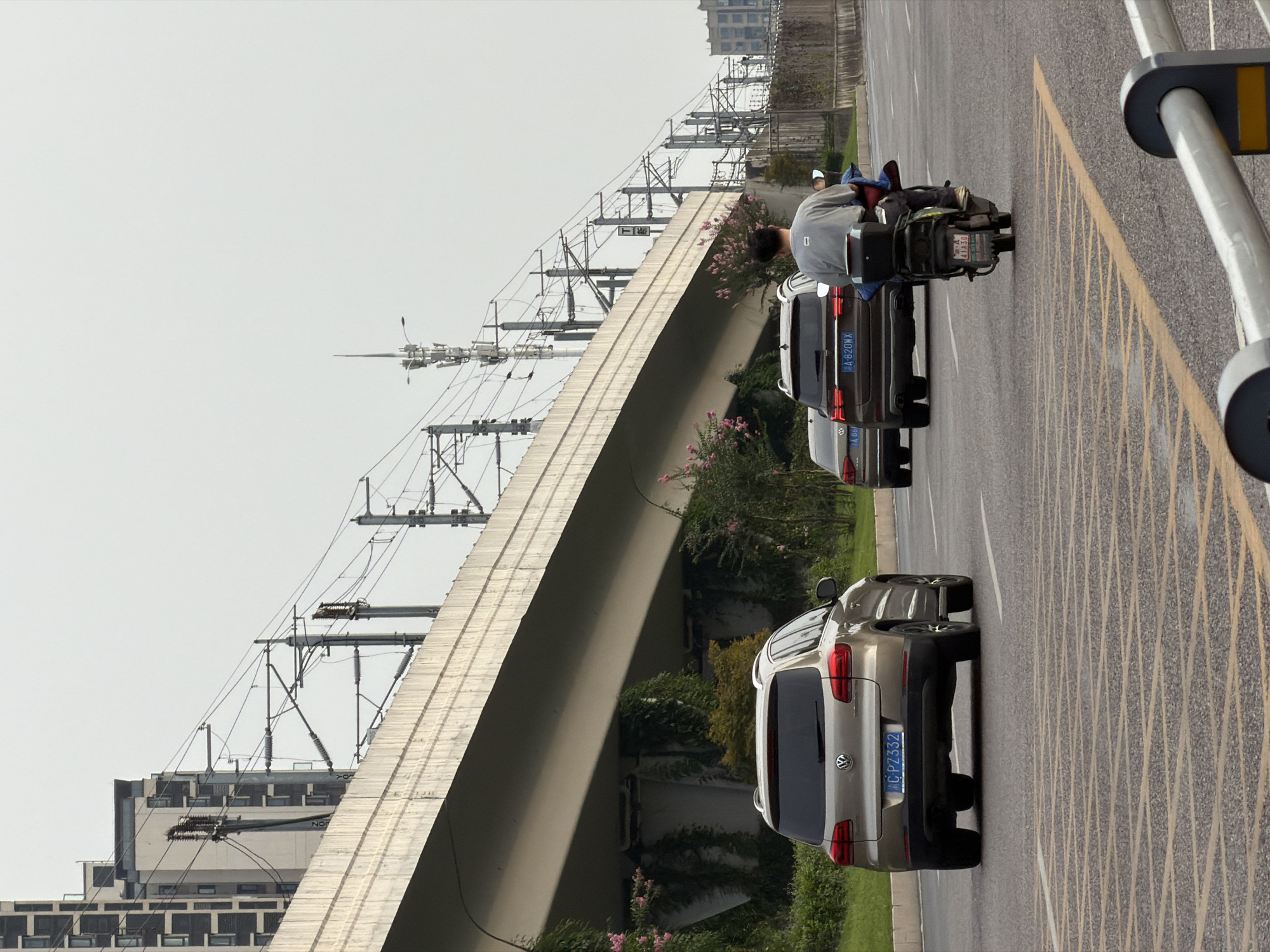 This image depicts a street scene in Jiangjin, China, with a multi-level overpass dominating the background. The overpass supports what appears to be railway tracks, indicated by the numerous overhead electrical wires and support structures. Buildings are visible beyond the overpass, some with a modern architectural style featuring multiple windows.
In the foreground, a paved road with yellow markings is visible. Several vehicles are present: a silver Volkswagen Tiguan SUV is in the left lane, followed by a dark SUV in the right lane. A person is riding a scooter in the right lane, also heading away from the camera. The scooter rider is wearing a grey shirt and blue pants. The weather appears overcast. Vegetation, including some pink flowering bushes and ivy, grows along the sides of the road and under the overpass, suggesting a somewhat urban but green environment.