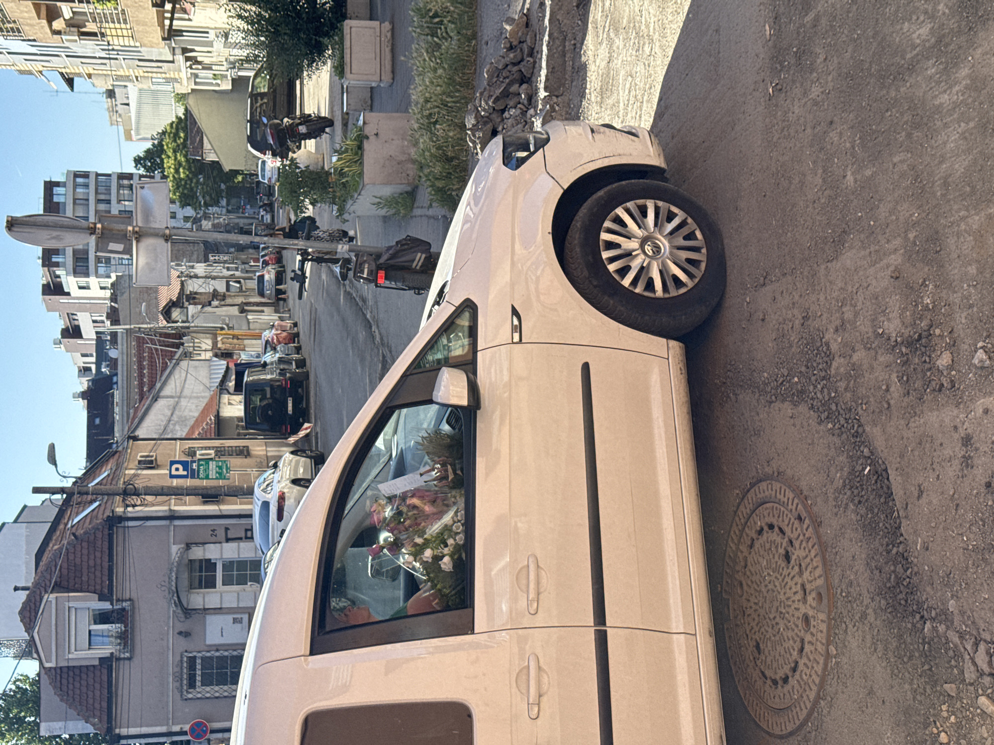 The image depicts a street scene in Vracar Urban Municipality, Serbia. A light-colored van, with flowers visible inside, is parked on the right side of the frame. To the left, the street stretches into the distance, revealing buildings of varying heights and architectural styles. Traffic is visible on the street. The foreground shows the curb and pavement, including a manhole cover. The street scene in the background includes a motorcycle, parked cars, and pedestrians. Buildings are visible on both sides of the street, with power lines strung between them. Street signs are also visible.