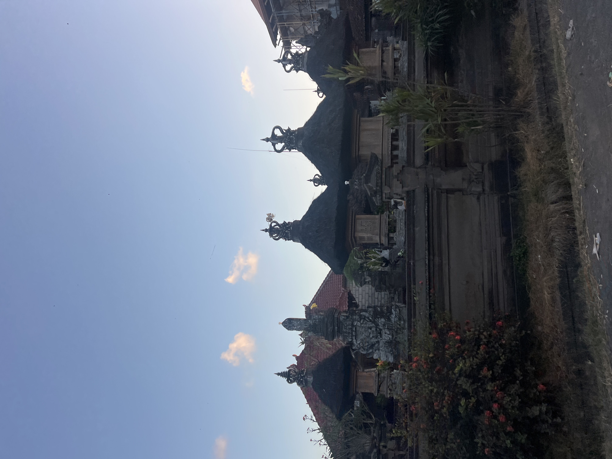 The image depicts a Balinese temple or shrine with its characteristic thatched roofs and ornate stone carvings. The structures are adorned with elaborate finials on top of the conical roofs. In the foreground, a bush with red flowers is visible, and a low stone wall runs along the front of the property. The background shows buildings with red tiled roofs and some construction scaffolding. The sky is a clear light blue with a few wispy clouds. The scene suggests an outdoor setting, likely in a rural or semi-rural area in Bali, Indonesia, given the architectural style. The specific location in the city of Kediri, Indonesia, cannot be confirmed from the image alone.
