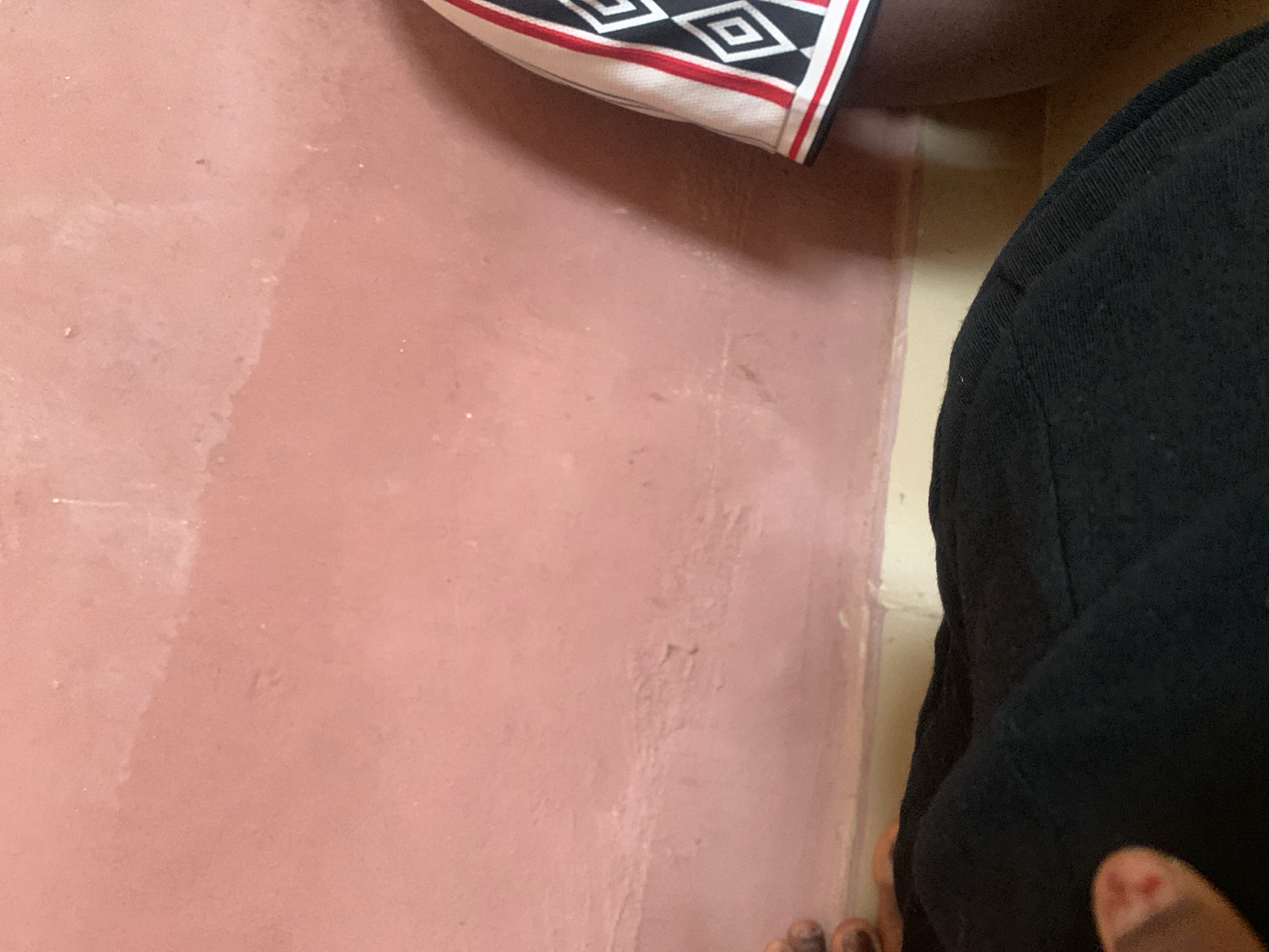 The image shows a close-up of a pink wall with some texture and discoloration. On the right side of the frame, a portion of clothing with a black, white, and red diamond pattern is visible. In the bottom left corner, a person's hand with what appears to be a small bandage or mark on one finger is visible, resting on black fabric, likely clothing. The scene is indoors, with the wall meeting a baseboard. The overall impression is that of a simple, everyday interior setting. The location of Jauro Yinu, Nigeria, is not determinable from the visual information in the image.