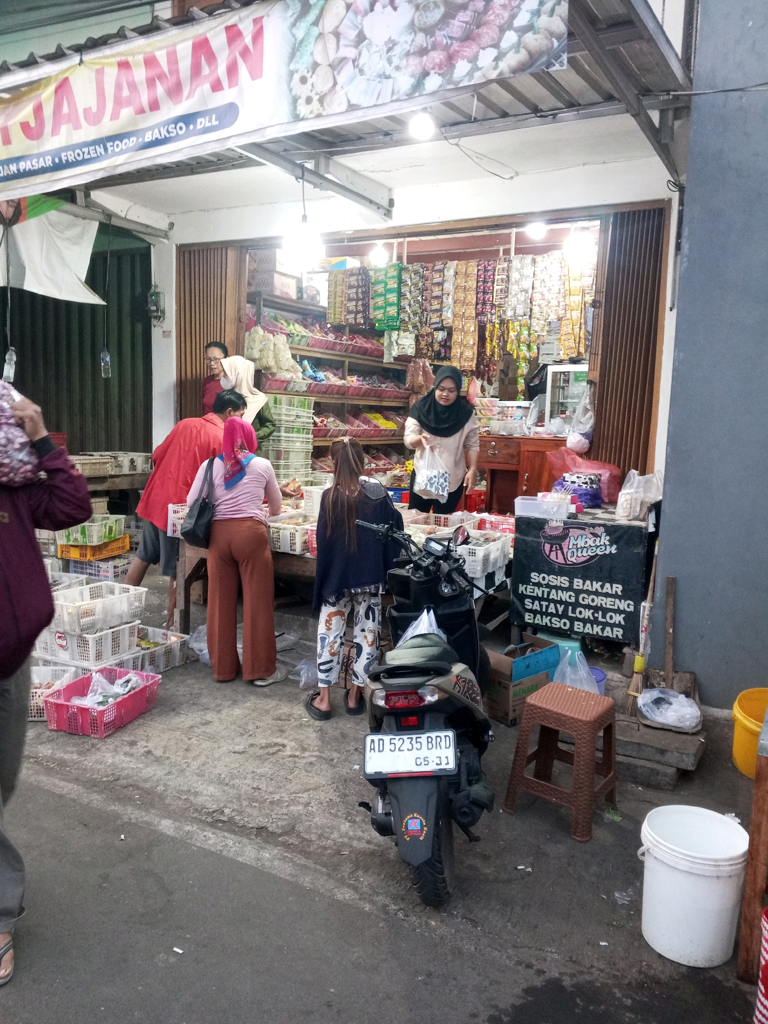 The image depicts an outdoor food stall in Gentan, Indonesia, likely on a street. A banner above the stall reads "YAYASAN" and below that "Jajan Pasar, Frozen Food, Bakso, DLL." Several people are present, appearing to be customers and vendors. In the foreground, a black motorcycle with a license plate reading "AD 5235 BRD 05-31" is parked. Various food items are displayed in baskets and on shelves within the stall. To the right, a sign advertises "Mbaak Queen" and lists food items like "Sosis Bakar," "Kentang Goreng," "Satey Lok-Lok," and "Bakso Bakar." The environment appears to be a bustling market setting with concrete ground and some debris.
