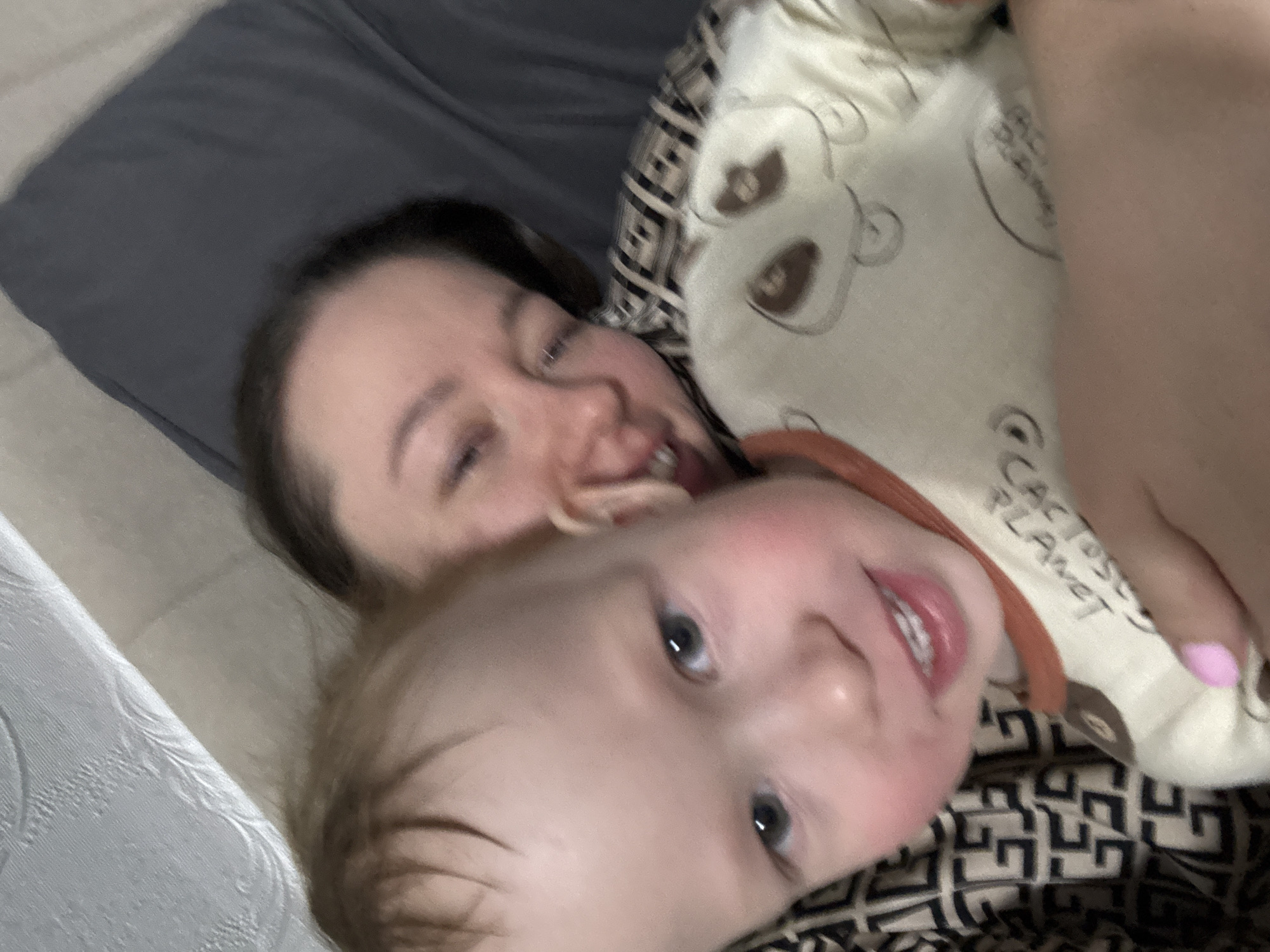 A woman and child are lying on a bed. The woman is in the background and the child is in the foreground, lying on top of her. The child is wearing a patterned white and brown shirt with an orange collar that has the words "CACTOSE PLANET" printed on it. A hand with pink nail polish is holding the child. The location is Ферма, Russia.