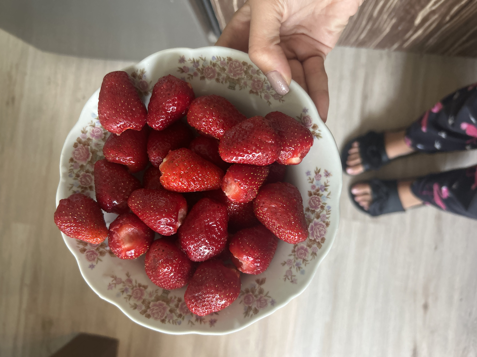 A top-down, eye-level shot features a bowl full of strawberries being held by a hand. The bowl is white with a floral pattern around the rim. The strawberries are red and appear to be ripe. The hand holding the bowl has a light skin tone and polished fingernails. In the background, parts of a person's legs and feet are visible, wearing black sandals and patterned pants. The floor has a light wooden finish. The scene appears to be indoors, possibly in a home kitchen, although the specific city and location are unknown.
