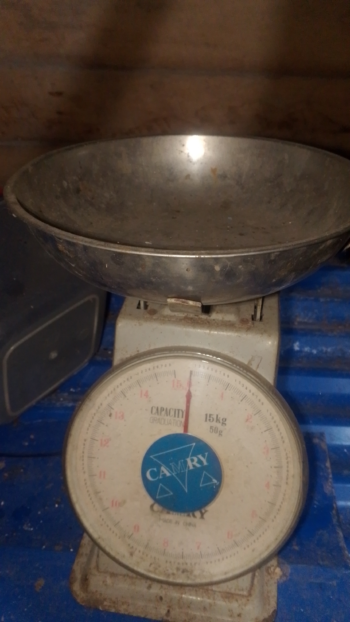The image shows an old, possibly vintage, mechanical scale with a large metal pan on top. The scale has a circular dial with markings for weight measurements in kilograms, with a needle pointing towards approximately 15 kg. A blue circular sticker with the brand name "CAMRY" is affixed to the dial. The scale appears to be dusty and slightly worn, suggesting it may be used or stored in a less than pristine environment. The background consists of what appears to be a wooden surface above and a blue corrugated surface below. The overall impression is of an object found in a workshop, storage area, or a residential setting in Olak-Olak Kubu, Indonesia. There are no people or any indication of recent activity.