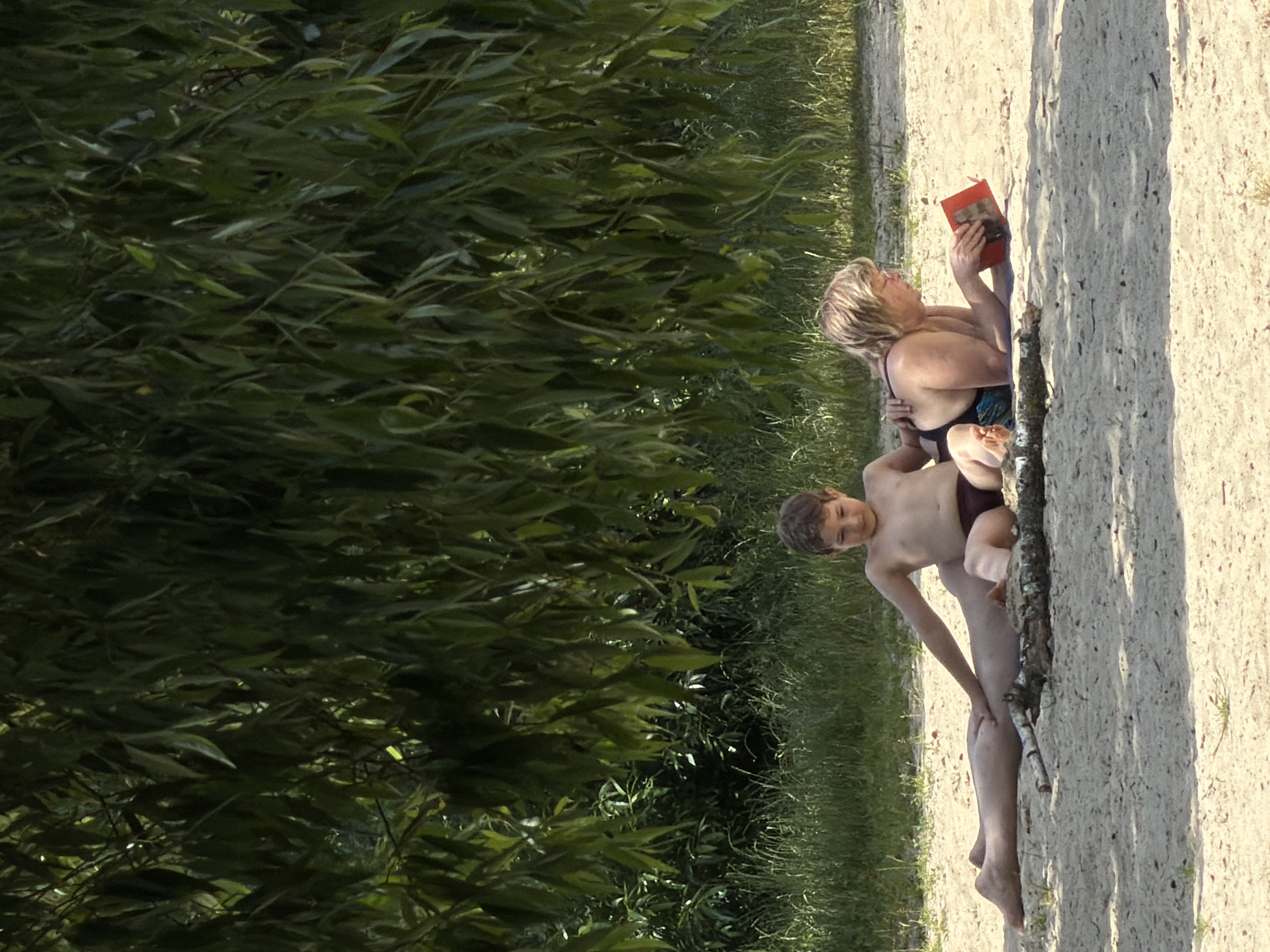 In this vertical, eye-level medium shot, two people are seen from the torso up, sitting on a sandy bank beside lush green foliage. A woman with short, blonde hair is on the left, wearing a dark swimsuit. She is holding a red book with a picture on its cover, reading it with a serene expression. Next to her, to the right, is a young boy, also in swimwear, appearing to be nestled into her side. His gaze is directed slightly upwards, and his expression is placid.
The setting appears to be a sunlit outdoor area, likely a beach or a lakeshore given the sandy ground and dense vegetation. The sun casts shadows from the foliage onto the sand, suggesting it is daytime, possibly late morning or early afternoon given the strong sunlight. The overall mood is one of peaceful relaxation and quiet companionship. No visible text is present in the image.