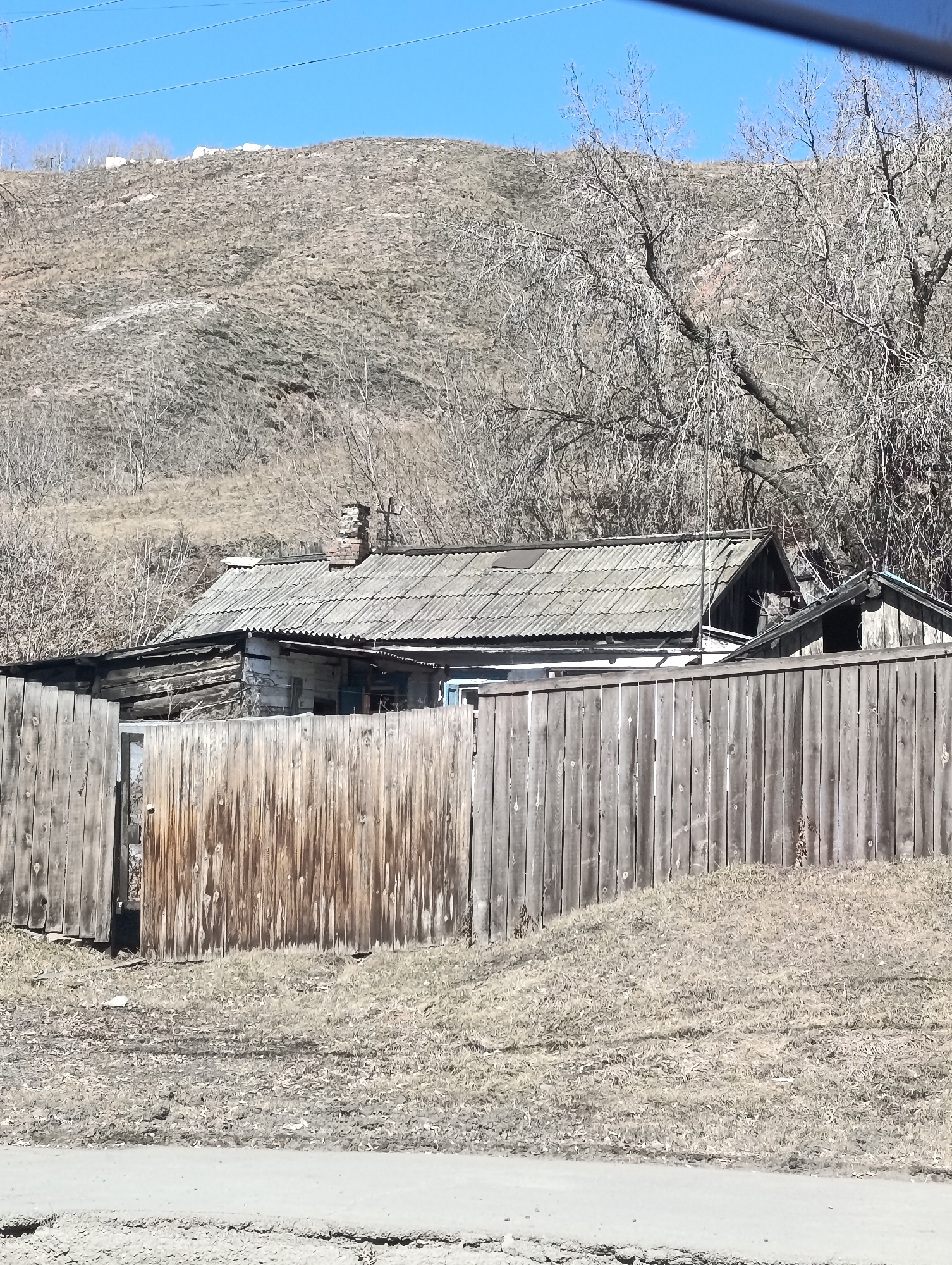 A daytime shot in what appears to be a rural or economically depressed area, potentially near Krasnoyarsk, Russia, shows an old, dilapidated wooden house behind a weathered wooden fence. The house has a grey, corrugated roof and a brick chimney. The surrounding area is covered in dry grass and vegetation. A hill is visible in the background, and bare trees are scattered throughout the scene. A road is partially visible at the bottom of the image. There are no people visible.