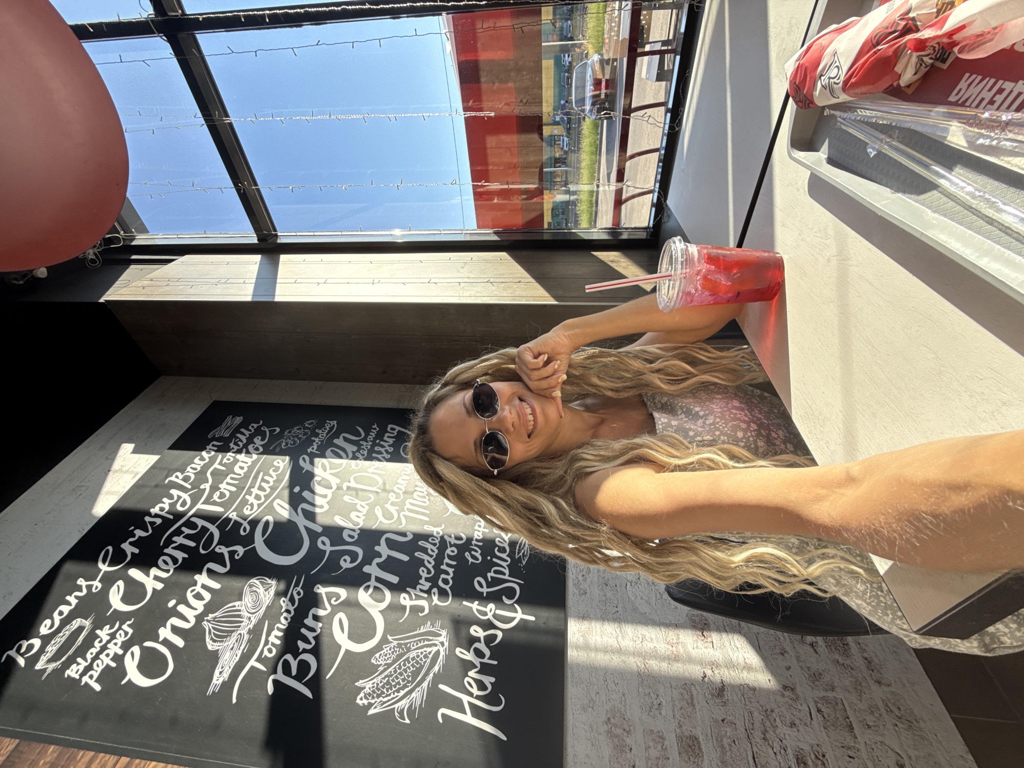 A woman with long, blonde, wavy hair and sunglasses is seated at a table in a cafe. She is smiling and resting her chin on her hand. In front of her is a red drink in a clear plastic cup with a straw. A food tray with utensils and a wrapped item is also on the table. Behind her is a chalkboard menu with various food items written in white chalk, including "Beans," "Crispy Bacon," "Tortilla," "Black Pepper," "Onions," "Cherry Tomatoes," "Lettuce," "Chicken," "Tomato," "Buns," "Corn," "Salad Dressing," "Shredded Carrot," "Wrap," and "Herbs & Spices." The cafe has large windows that offer a view of a street with buildings and cars. The overall lighting suggests it is daytime, with sunlight casting shadows. The scene is likely located in the city of Rusanovo, Russia, given the context.