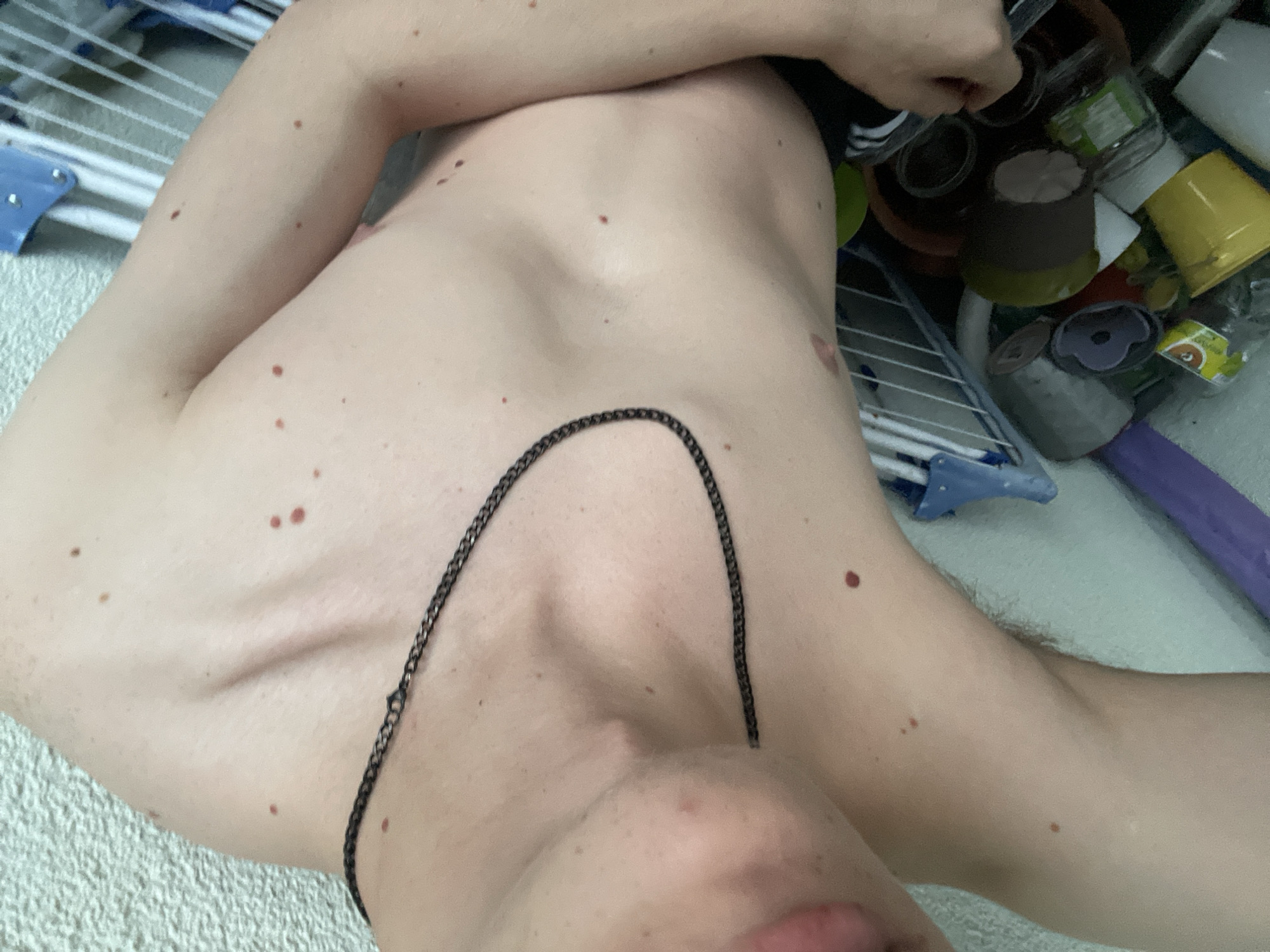 This media file is a close-up, overhead shot of a person's bare chest and upper arm, adorned with a dark chain necklace. The individual appears to be lying down, with their body positioned diagonally across the frame. The skin is fair and marked with several moles of varying sizes and shades of brown, including a prominent cluster of three moles on the collarbone area.
The background is cluttered with various objects, suggesting an indoor setting. Visible items include a blue drying rack with white bars, a collection of assorted containers (some appearing to be plastic jars or bottles, possibly holding cleaning supplies or food items), a brown cylindrical object, and a purple cylindrical item. The overall impression is one of a domestic or utility space, perhaps a laundry room or a storage area, with items haphazardly arranged.
No specific activity is clearly depicted, other than the subject's passive presence in the scene. The lighting appears to be artificial, with no clear indication of natural light sources. The time of day and weather conditions are indeterminate. There is no visible text within the frame. The image focuses on the texture and details of the skin and the surrounding objects, creating an intimate but somewhat unspecific portrayal.