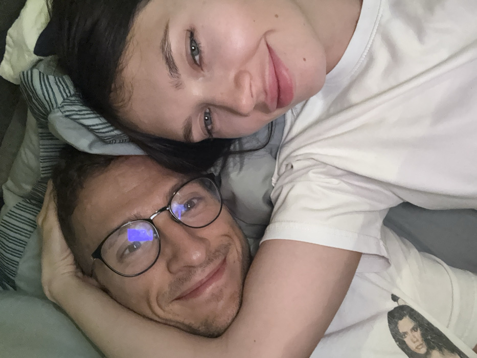 The image depicts two individuals, a male and a female, situated in what appears to be a bed, likely in a residential setting in Herzliya, Israel. The perspective is an overhead shot looking down at the subjects.
The male is positioned on the left side of the frame. He has short, dark hair, wears dark-rimmed eyeglasses with visible reflections, and a light-colored beard. He is wearing a white t-shirt with a dark graphic print visible on his left shoulder. His left arm is bent, with his hand resting behind the head of the female subject, suggesting an embrace or close proximity. He is smiling towards the camera.
The female is positioned on the right side of the frame. She has long, dark hair and is wearing a plain white t-shirt. She is also smiling towards the camera.
Both individuals are lying on what appears to be pillows and bedding with a light blue and white striped pattern visible in the upper background. The lighting is soft, originating from an unseen source, creating reflections in the male's eyeglasses.