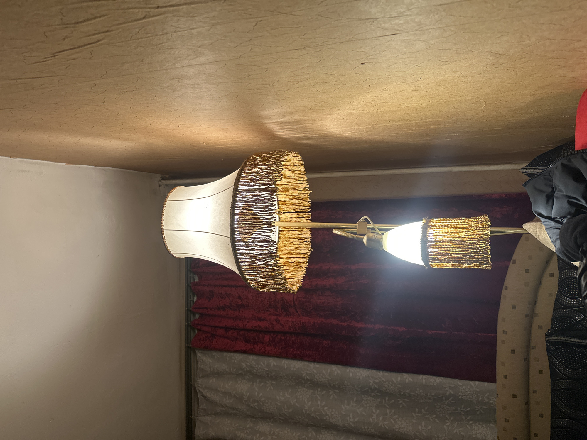 The image shows an indoor scene. In the foreground is a vintage-style lamp with two shades, one larger and one smaller, both featuring fringe detailing. Behind the lamp, there are curtains, with the top layer in a dark, possibly maroon or burgundy, plush material, and the layer below in a lighter, patterned fabric. In the background, there are clothing items and a wall painted in a neutral tone. The ceiling is also a neutral color with a textured finish. The image is taken at an angle, and the light from the lamp is bright, suggesting it's illuminated. I cannot infer the location of Sofia, Bulgaria from the image.