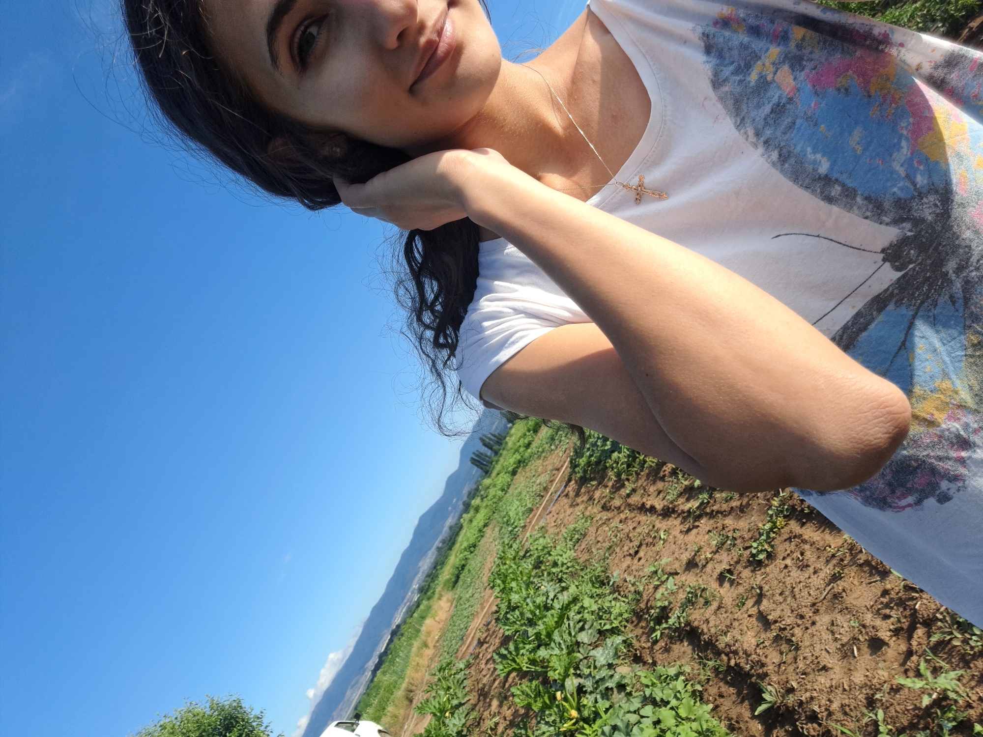The media file is a close-up, angled selfie of a young woman outdoors. She has dark hair, and her face is partially obscured as she rests her head on her hand, with her arm bent. She is wearing a white t-shirt with a large, colorful butterfly graphic on it and a delicate cross necklace. The background shows a bright, clear blue sky, a field of green plants, and distant hills or mountains. The sunlight suggests it is daytime, likely morning or afternoon. The scene appears to be rural, possibly a farm or garden. The woman's expression seems relaxed and content. There is no visible text in the media file.