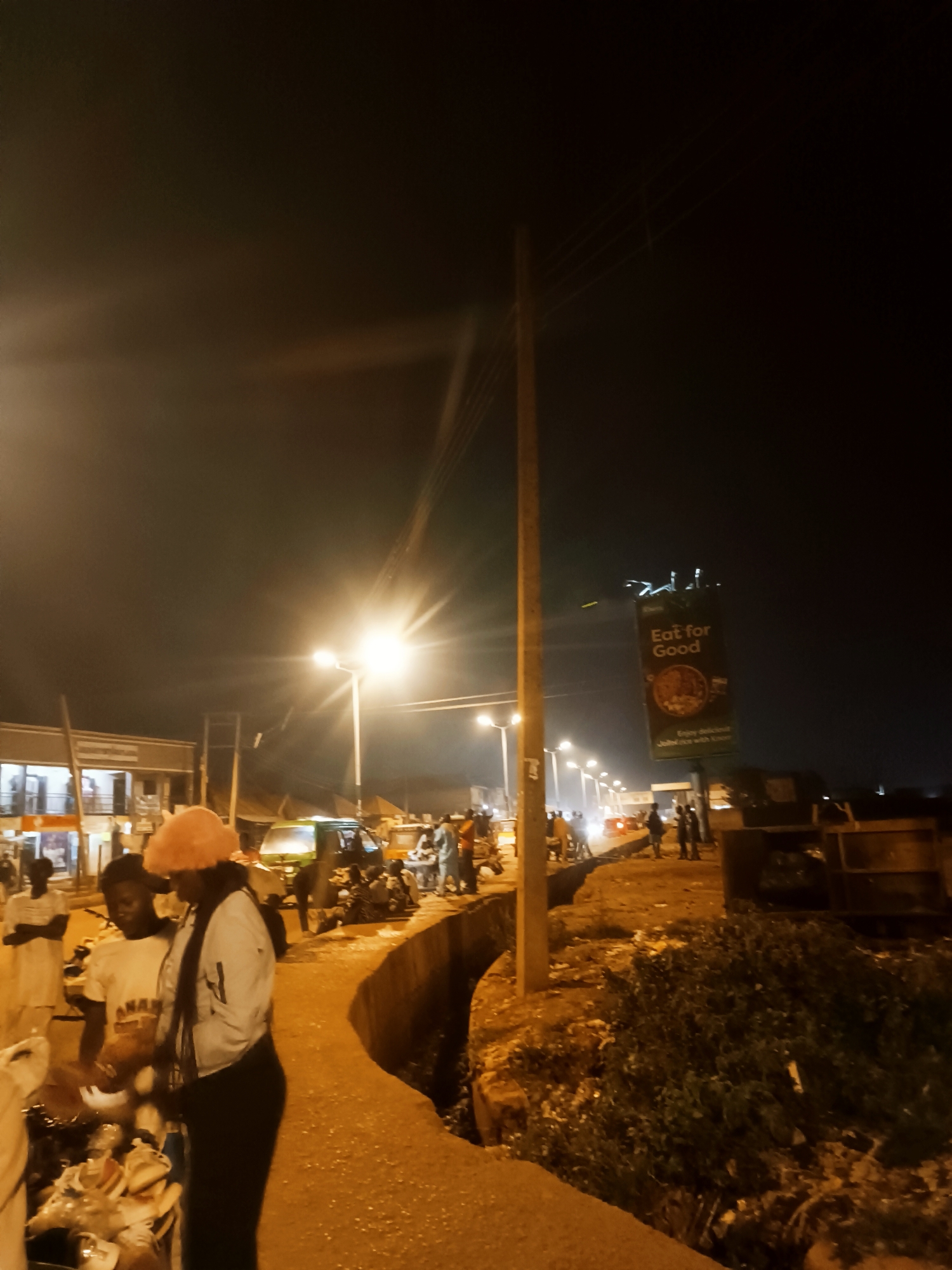 The image captures a night scene in Bukuru, Nigeria. A street illuminated by bright streetlights is bustling with activity. Several people are visible, including a woman wearing a pink hat and a light-colored jacket who is standing near a vendor's stall, possibly examining goods.
The environment is slightly rough, with a concrete barrier separating a walkway from a ditch filled with vegetation and some trash. In the background, there is a billboard advertising "Eat for Good" jollof rice. A variety of vehicles, including motorcycles and auto rickshaws (tricycles), can be seen on the road. The overall setting is urban, with buildings visible in the background under the night sky.