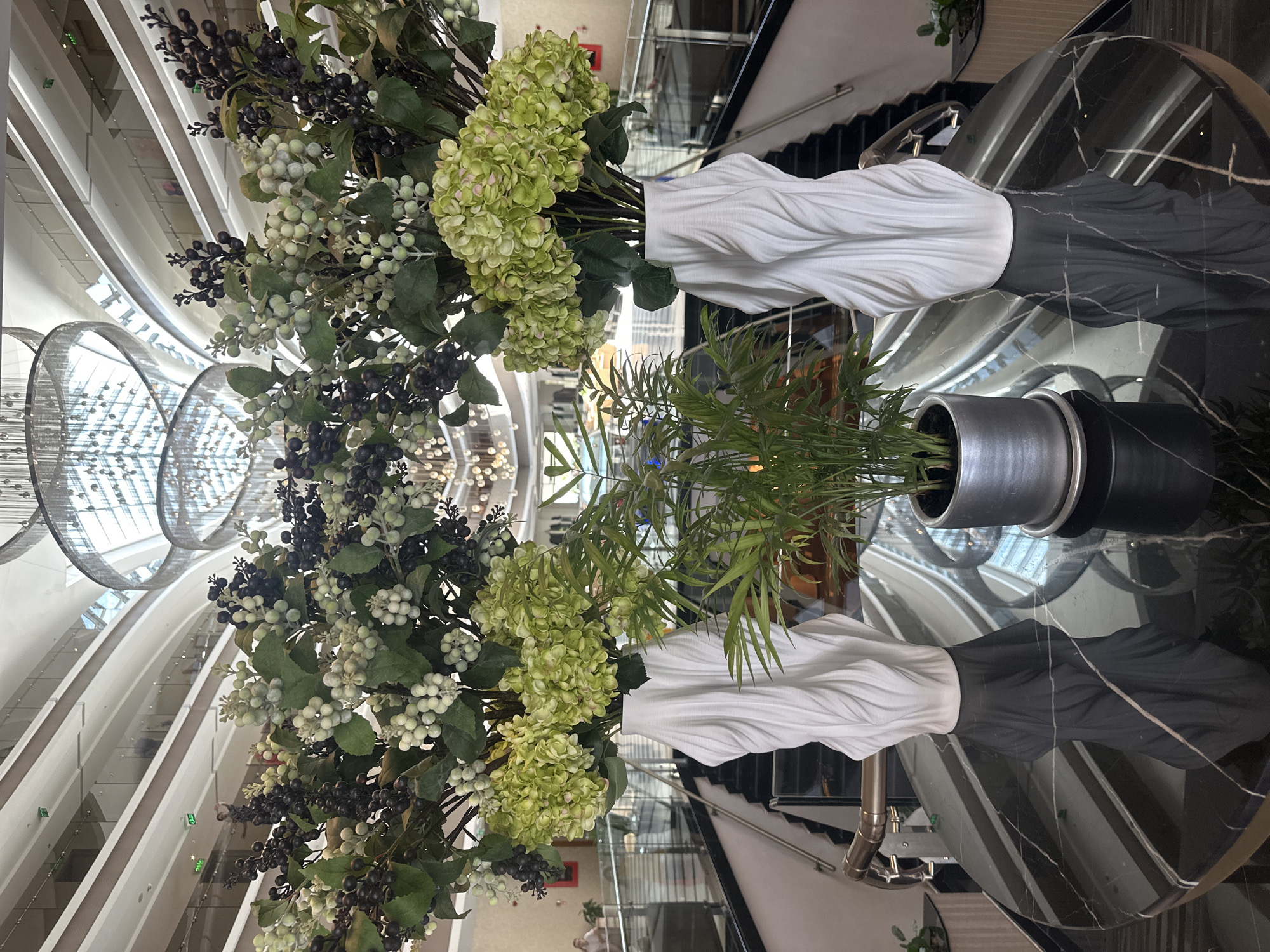 This is a photograph taken indoors, likely in the lobby of a hotel or resort, with a focus on decorative floral arrangements and interior architecture. The scene is set on a large, circular, polished black marble table with white veining.
On the table are two tall, white, sculpted vases with a draped fabric-like appearance. Each vase holds a large arrangement of artificial flowers and berries. The arrangement on the left features clusters of light green hydrangeas and darker green foliage, interspersed with bunches of dark purple and light green berries. The arrangement on the right appears similar, though the proportions of berries and flowers may differ slightly.
In the center of the table, between the two large arrangements, is a smaller potted plant with feathery green leaves. This plant is housed in a cylindrical metallic grey pot with a visible saucer.
The background of the image reveals a grand, multi-story interior space with a distinctive architectural style. Curved balconies with glass railings overlook a central atrium. A spiral staircase with a metallic handrail is visible on the left side of the frame, ascending from a lower level. Above, a large, modern chandelier with multiple tiers of shimmering circular elements hangs from the ceiling, reflecting the ambient light. The overall impression is one of spaciousness and contemporary design.
While the image is set in Manavgat, Turkey, the specific location within the city is not identifiable from the visual information presented. It is likely a luxury hotel, resort, or a commercial building designed for high-end clientele. No people are clearly identifiable in the immediate foreground or central to the composition, although indistinct figures can be seen on the balconies in the background. The lighting appears to be a combination of ambient natural light and artificial interior lighting, creating a bright and well-lit environment.