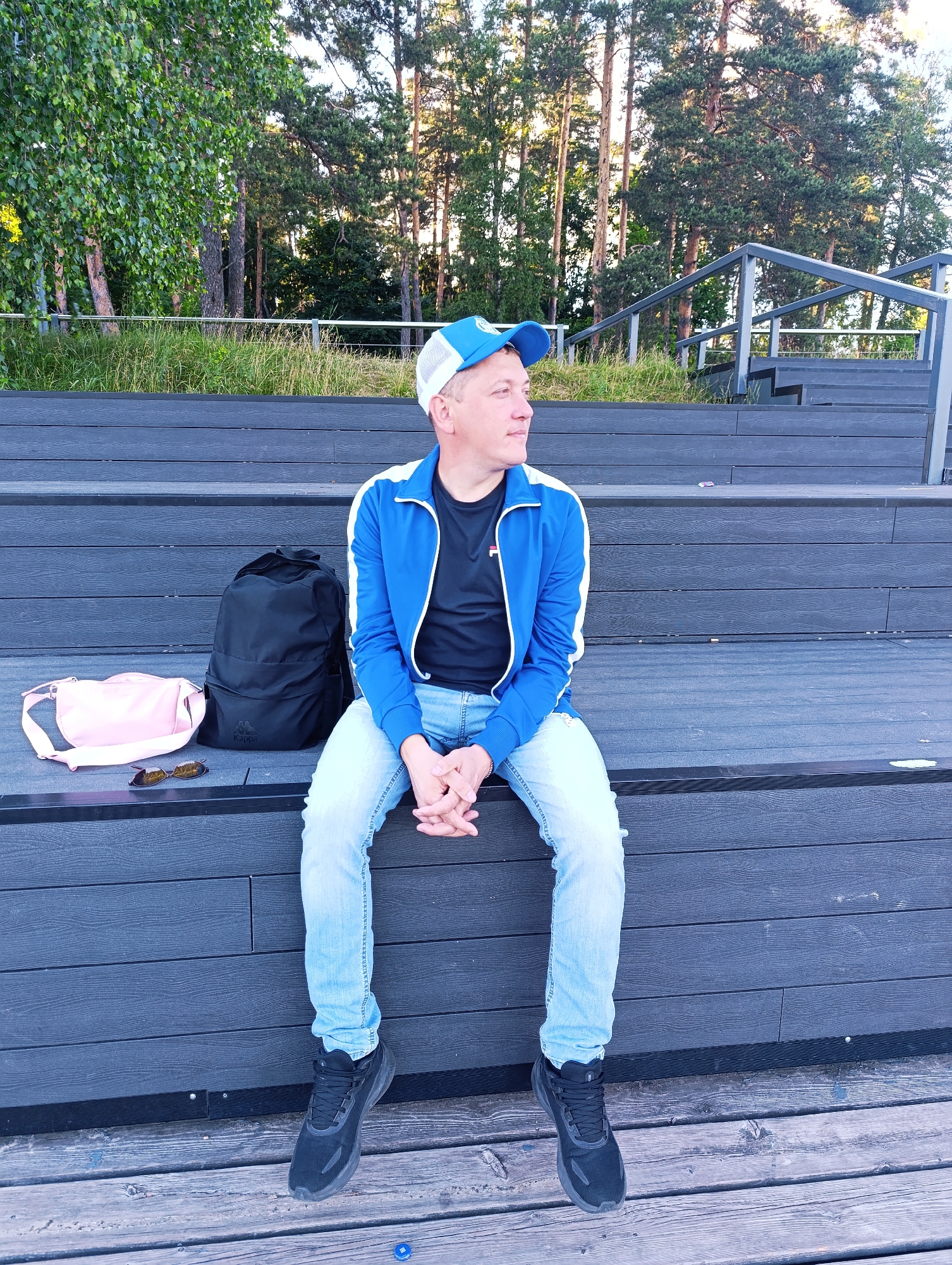 A man wearing a blue and white baseball cap, a blue and white jacket, a black t-shirt, and light blue jeans sits on dark wooden bleachers. He is looking to his right with his hands clasped. To his left on the bleachers are a black backpack, a pink crossbody bag, and a pair of sunglasses. The scene is outdoors, with a background of trees and a clear sky, suggesting it is daytime. The man appears relaxed. The text "Kappa" is visible on the black backpack. The location is Chernogolovka, Russia.