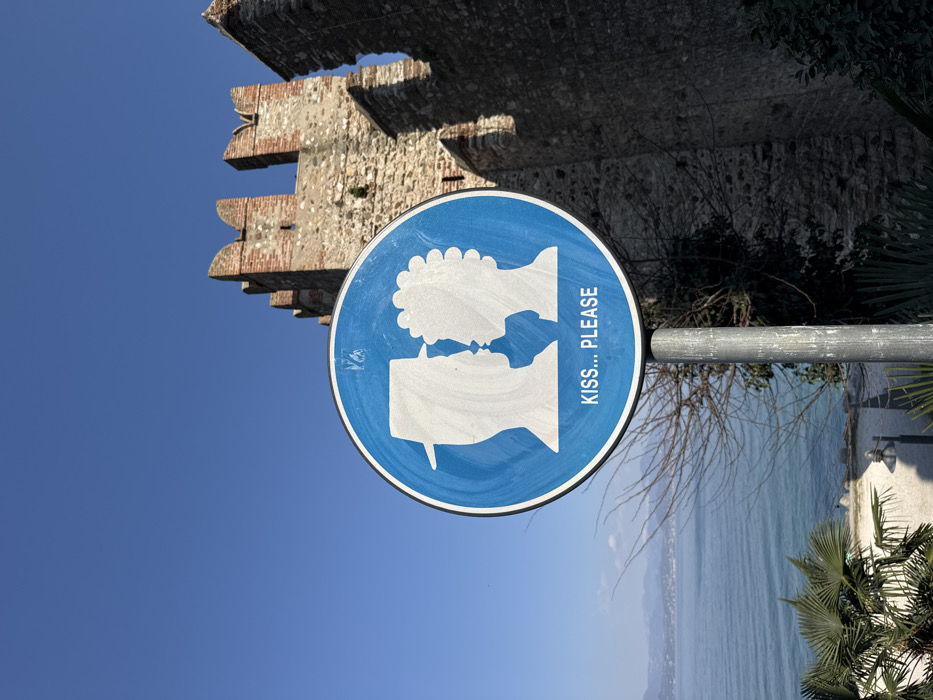 This image captures a circular blue and white instructional sign mounted on a grey metal pole, situated in Sirmione, Italy. The sign, showing minor surface wear, features two white silhouettes on a blue background: a left profile of an individual wearing a fedora-style hat facing a right profile of an individual with textured, possibly curly, hair. Below the silhouettes, white text reads "KISS... PLEASE". To the immediate right and extending into the background is a rough-hewn stone wall with red brick crenellated battlements, characteristic of the Scaligero Castle. To the left, a vast expanse of calm, blue water, identified as Lake Garda, stretches towards a distant, hazy mountain range under a clear, azure sky. In the lower foreground, surrounding the sign's base, sparse, deciduous branches intertwine with green, broad-leafed plants, possibly palms. A paved pathway and a dark-colored streetlamp are partially visible near the lake's edge. No individuals are physically present in the scene, though the sign implies an action.
