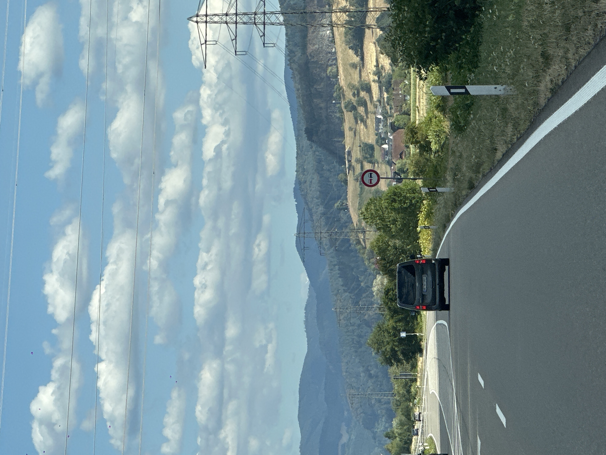 The image captures a scene on a highway, likely in the vicinity of Biberach, Germany, based on the signage and landscape. The perspective is from inside a vehicle, looking forward. A dark-colored minivan is driving in the right lane, ahead of the viewer. The road is paved asphalt with white lane markings. To the right of the road, there is a grassy verge with wildflowers and several white marker posts. A circular traffic sign with two red horizontal bars indicating a no-overtaking zone is visible.
The background features rolling hills covered in trees and fields, interspersed with houses of a small village. Several large electricity pylons with power lines stretch across the landscape. The sky is partly cloudy, with fluffy white clouds against a blue backdrop. The overall impression is a sunny day in a semi-rural setting.