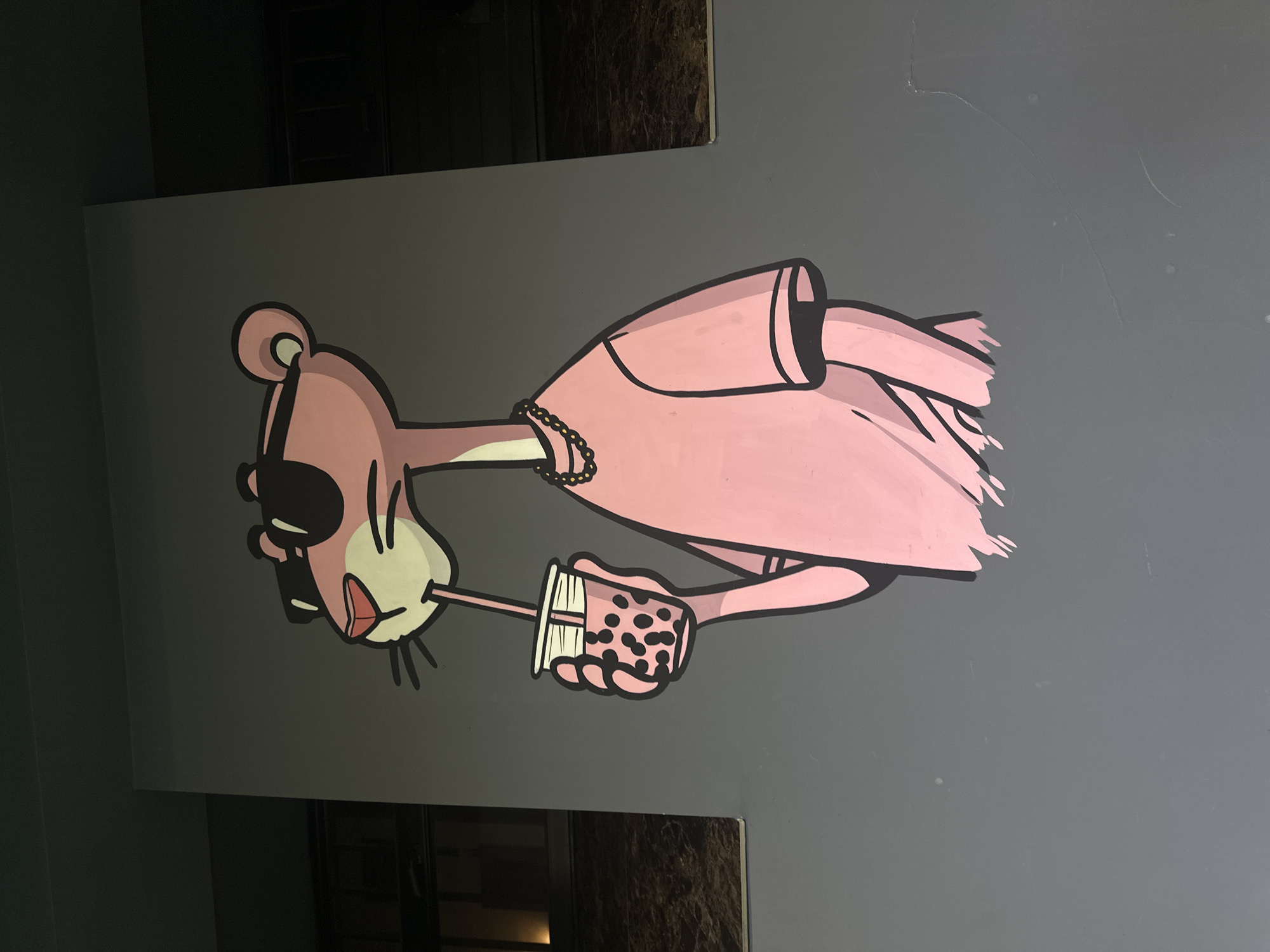A mural in Yerevan, Armenia, depicts a pink panther-like cartoon character wearing sunglasses and a pink dress, sipping from a bubble tea cup. The mural is painted on a gray wall.