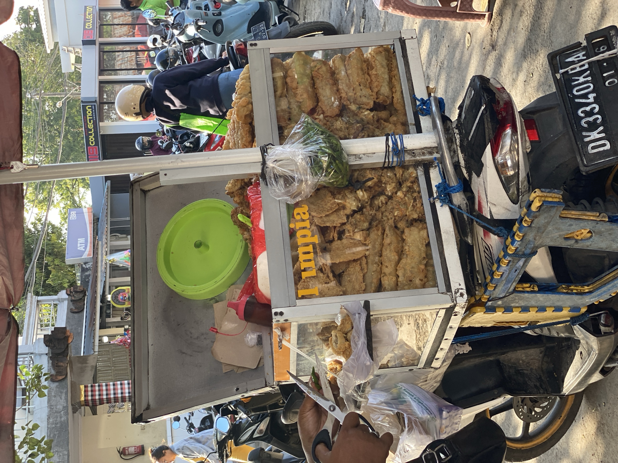 A street vendor in Singakerta, Indonesia, is selling food from a cart attached to a motorcycle. The cart contains fried snacks, including tempeh, displayed in three glass-fronted tiers. The vendor is using scissors to prepare a plastic bag containing food for a customer. In the background, other motorcycles are parked on the side of the road, and shopfronts are visible. A license plate reading "DK 3340 KBA" is attached to the vendor's motorcycle.