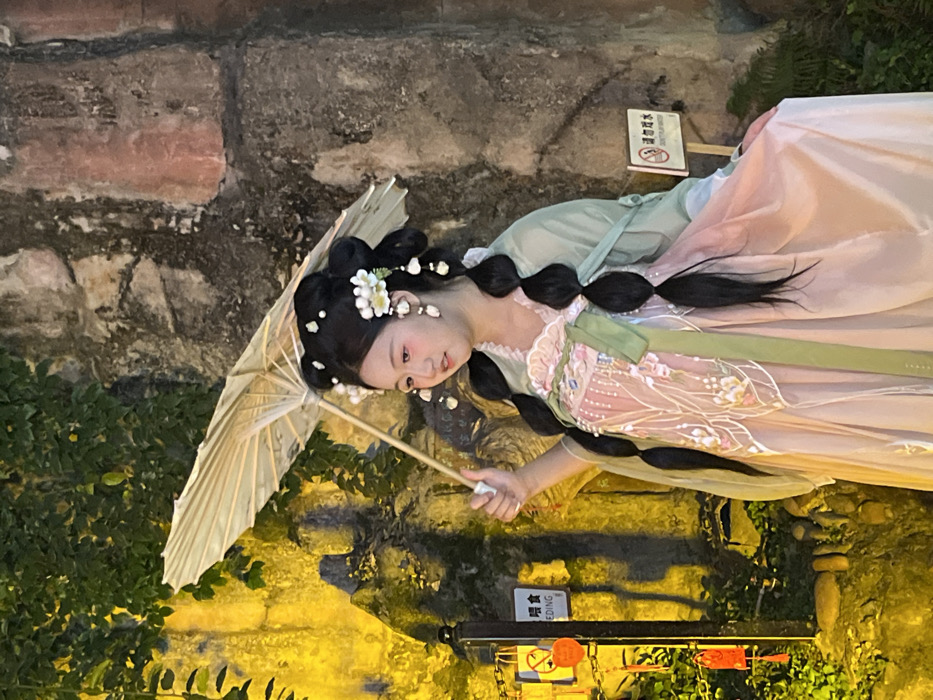 The image displays a young East Asian female standing outdoors at night in Yuzhong District, China. She is positioned slightly right of the frame's center, facing left with a subtle expression. She wears a traditional Chinese-style costume consisting of a light pink, long dress with intricate white floral embroidery on the bodice and upper skirt, a wide light green sash, and a sheer, light green overgarment. Her black hair is styled in an elaborate manner, featuring multiple buns adorned with white flowers and white beaded strands, with two prominent, long, thick braids extending down her front. She holds an open, light yellow traditional umbrella in her raised right hand.
The background features textured stone walls. The left portion of the wall is covered with dark green foliage, brightly illuminated by a strong yellow light source. A dark pole on the left displays several orange heart-shaped ornaments and a white sign with partial visible English text "EEDING" and a "No Smoking" symbol. The right portion of the wall exhibits a darker, reddish-brown tone. A white rectangular sign with a "No Smoking" symbol and corresponding Chinese characters is affixed to the wall near the bottom right. The scene is artificially lit, creating contrasts between illuminated areas and shadows.