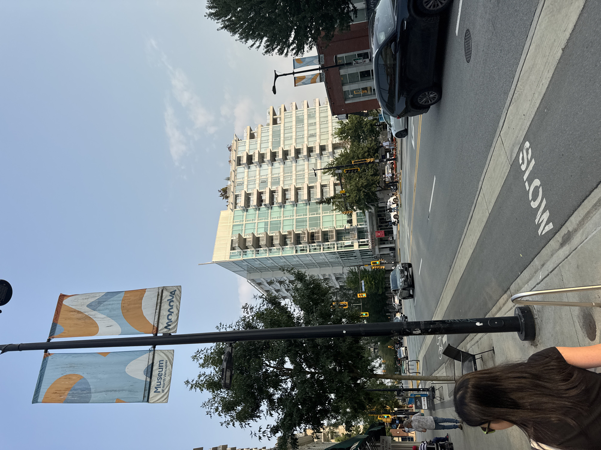The image captures a street scene in the city of North Vancouver, Canada. A modern, multi-story building with balconies and a curved facade dominates the background. In the foreground, a sidewalk with a tree and a lamppost is visible. Two banners hang from the lamppost, one displaying "Museum of North Vancouver" and the other with abstract orange and blue patterns. A black SUV is driving on the street, and other vehicles are present in the distance. A person is walking on the sidewalk on the left side of the image, and another person is seen from behind in the lower left corner. Traffic signals are visible on poles along the street. The sky is clear with some light clouds.
