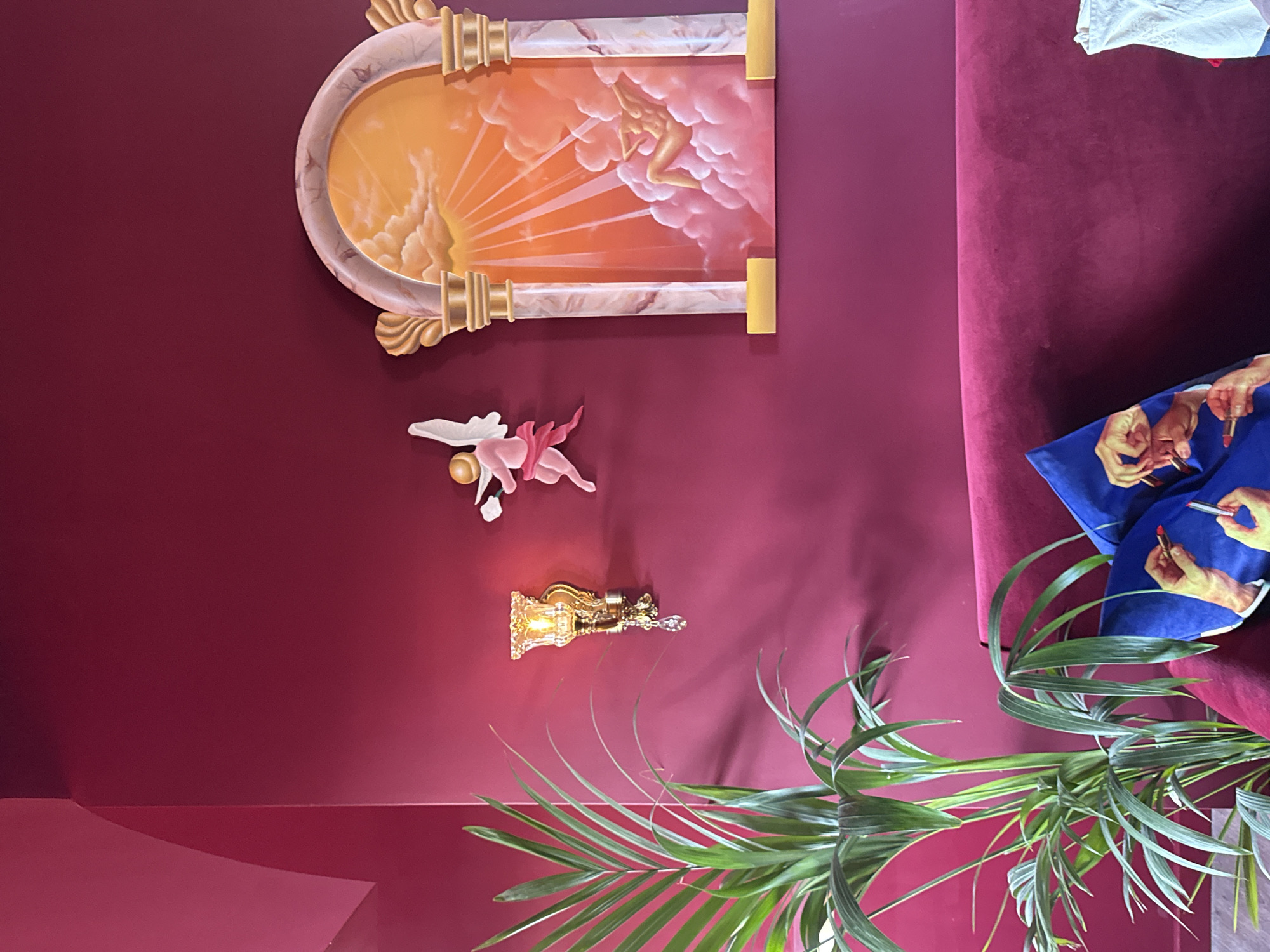 This image depicts a highly decorated interior room at The Mandrake Hotel, located in London, United Kingdom. The environment is characterized by deep magenta walls and a plush, matching velvet sofa occupying the lower portion of the frame.
On the left side of the image, a tall, leafy green palm plant stands, its fronds extending upwards. Above and to the right of the plant, an ornate golden wall sconce is mounted, featuring a decorative glass shade that emits a warm, ambient light, and a single dangling crystal. Further to the right, a pink three-dimensional relief sculpture of a winged cherub, positioned mid-flight, is affixed to the wall.
Dominating the right side of the wall is a large decorative art piece resembling an arched architectural niche or window. It features two marbled columns with golden capitals and bases, framing a painted scene. This painting depicts a vibrant orange and pink sky filled with clouds, radiating sunbeams, and a reclined nude figure, possibly female, amidst the clouds.
On the deep magenta velvet sofa in the foreground, a blue throw pillow with a pop-art style print is visible, showcasing multiple hands holding red lipsticks. To the far right of the sofa, a crumpled white fabric object rests. No human figures are present in the immediate scene, apart from the depicted subjects in the artwork.