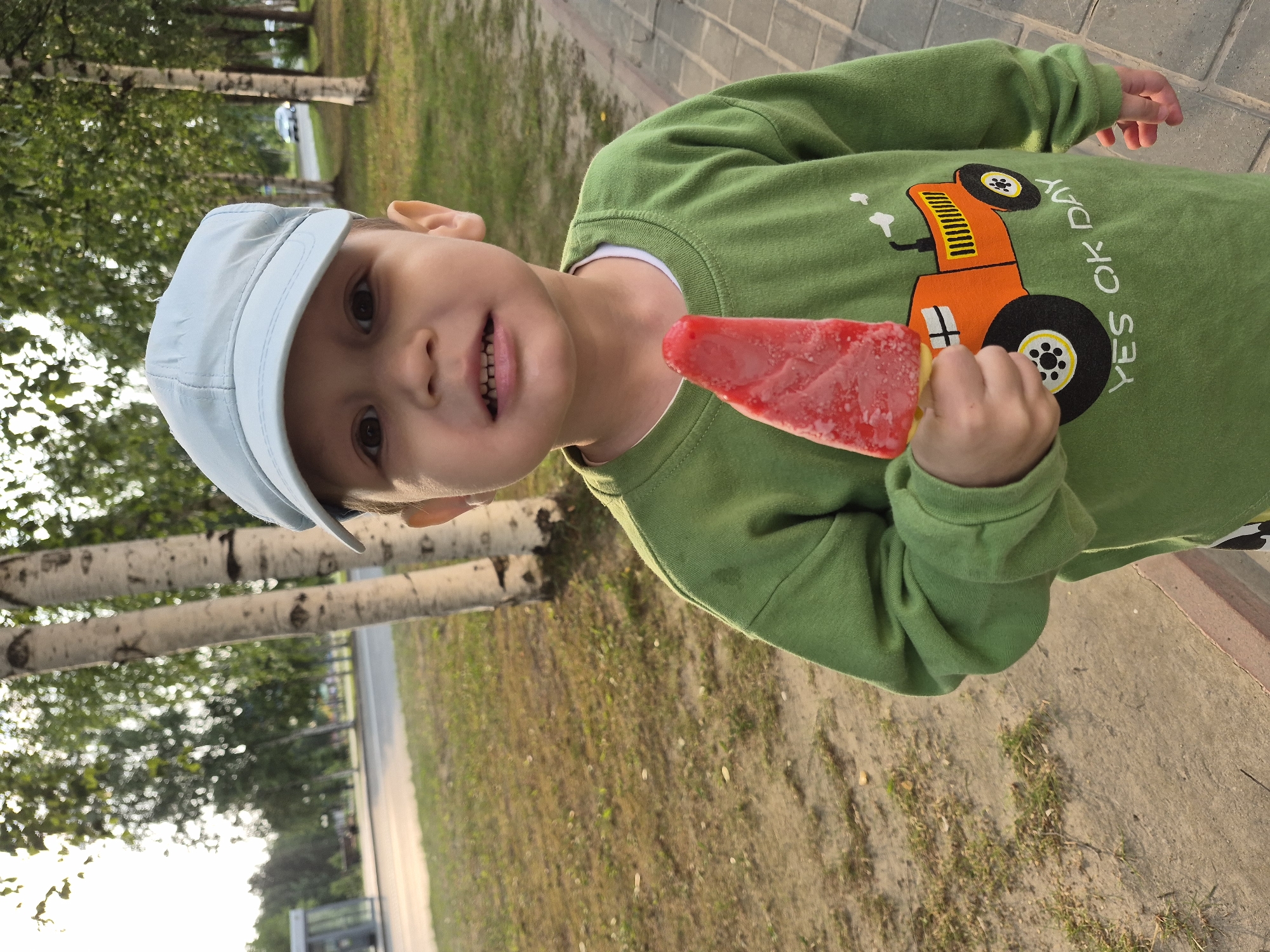 A young boy, wearing a light blue cap and a green long-sleeved shirt with a graphic of an orange truck that reads "YES OK DAY", is holding a red popsicle. He is outdoors, with trees and a paved walkway in the background. The sun appears to be low in the sky, casting a warm light on the scene, suggesting it is either early morning or late afternoon. The setting looks like a park or a residential area with grass and trees. The boy's expression is neutral, and he is looking directly at the camera. The overall mood of the image is casual and summery.