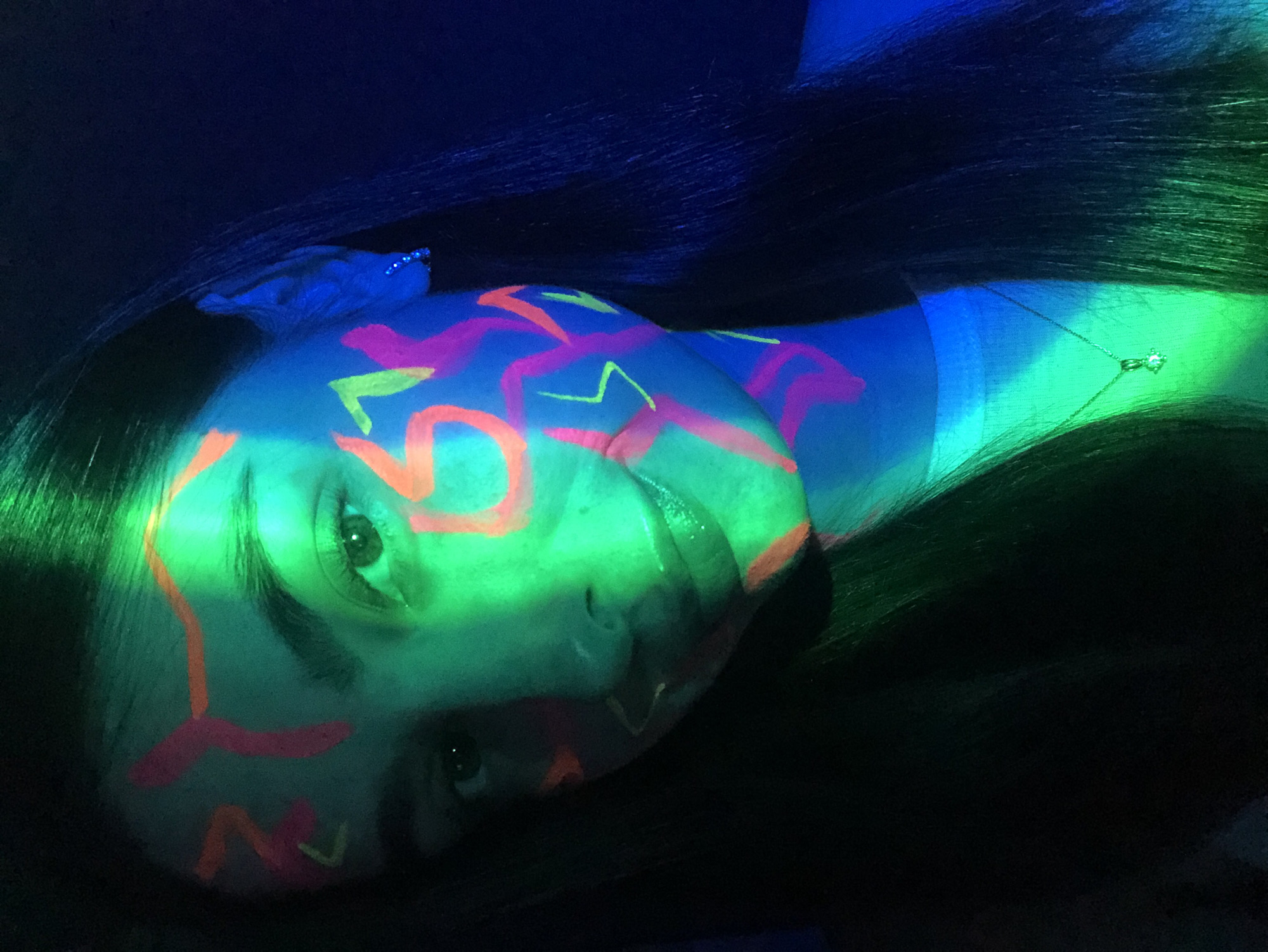 The image captures a person, likely female, lying or tilting their head horizontally in a dark, dimly lit environment under ultraviolet (UV) light. Their face, neck, and upper chest are adorned with intricate, abstract designs using fluorescent body paint in shades of neon pink, orange, and lime green/yellow. These paints glow intensely against the skin, which appears a pale blue-green under the UV light.
The person's gaze is directed towards the viewer, with one eye clearly visible and the other partially obscured by the angle. Their expression is neutral. Long, dark hair frames the face, appearing dark green where it catches the UV light and blending into the deep blue-black background elsewhere.
A delicate, silver-toned necklace with a small, sparkling pendant is visible on the person's neck, also reacting to the UV light. Part of a light blue or turquoise garment can be seen on the shoulder/upper arm area, glowing under the same light. The overall scene creates a vibrant, artistic, and somewhat otherworldly atmosphere, likely depicting an event or performance involving blacklight art or a themed party. There is no visible text in the image. The setting appears to be indoors at night, characterized by the absence of natural light and the exclusive use of UV illumination.