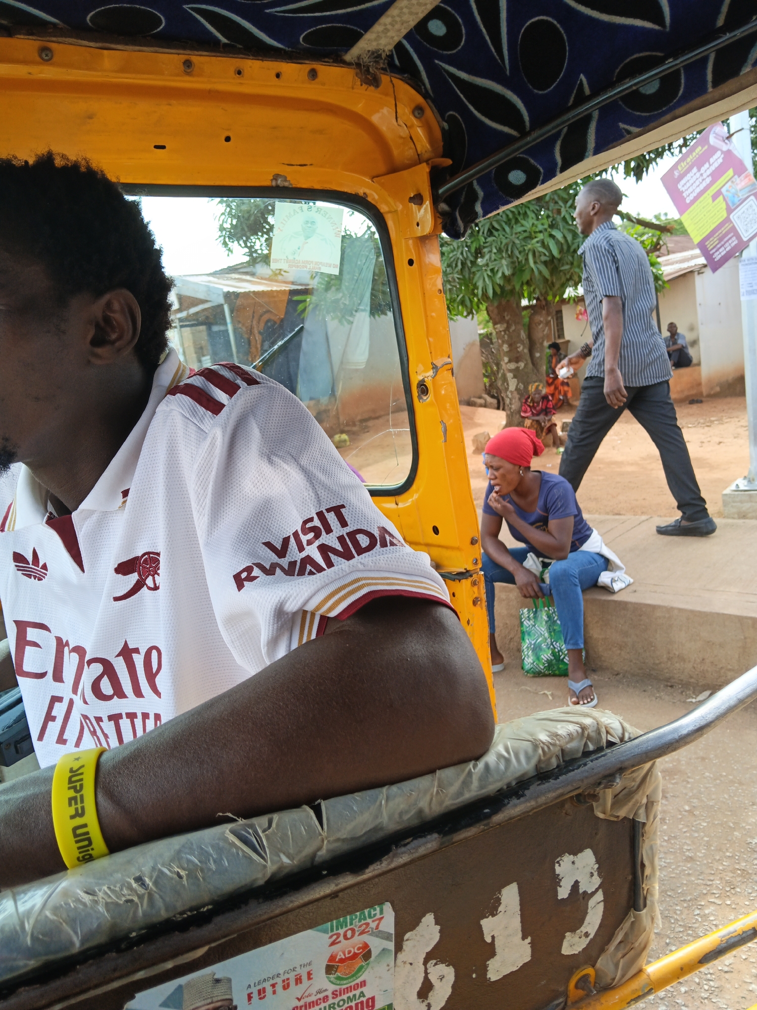 The image depicts an outdoor scene, possibly on a street in an urban environment, with a bright yellow auto-rickshaw prominently featured. A person, likely the driver, is seated in the foreground, facing left, wearing a white polo shirt with red trim and logos. Their arm is visible, adorned with a yellow wristband that reads "SUPER UNIQ." In the background, a man in a striped shirt and dark pants walks away from the viewer, holding a white object. To the right, a woman wearing a red headscarf and blue jeans is seated on a step, looking downwards and appearing to be eating. She has a green patterned bag beside her. Other individuals are visible in the background, suggesting a public space. The environment includes trees, buildings, and a dirt or paved ground. Several posters or advertisements are attached to poles and structures, one of which has "IMPACT 2027" visible. The overall impression is a candid snapshot of daily life in a bustling, possibly tropical, location.