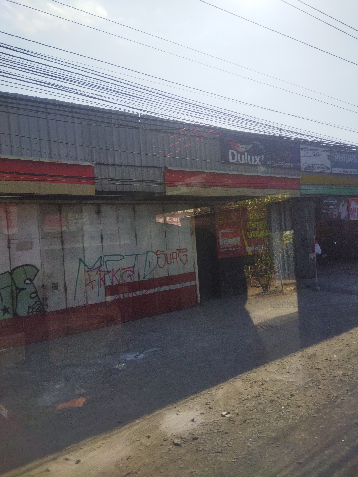 The image captures a street-level view of a commercial building in Nguter, Indonesia, under a bright, overcast sky. The building's facade is clad in corrugated metal, with horizontal red and yellow bands along the lower portion. A large, rolling metal door dominates the center of the image, showing signs of wear and featuring graffiti.
Above the door, a series of advertisements are visible. A prominent banner displays "Dulux let's colour," with a logo featuring a stylized bird. To the right, other advertisements include "GRAVI" and "PHILIPS," suggesting a retail or service establishment.
Reflected in the polished metal of the door are elements of the street and surrounding area, including a small tree, shadows, and what appears to be a red object on the ground. To the right of the main entrance, another section of the building is visible, with an open doorway revealing a glimpse of the interior. A red and white Indonesian flag is also visible, attached to a pole outside this entrance. The ground in front of the building is a mix of gravel and asphalt.