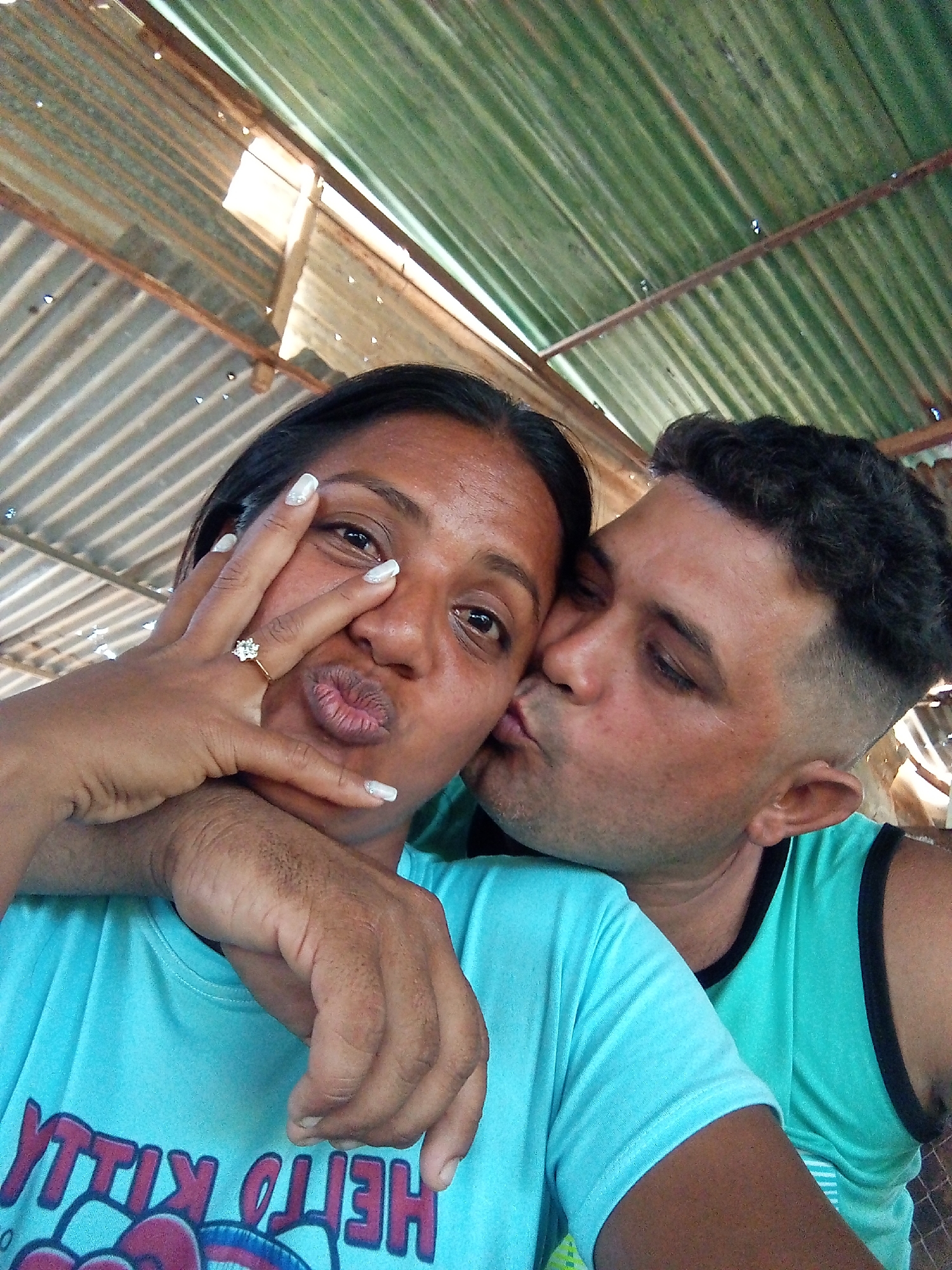 A medium shot captures a couple beneath a corrugated metal roof, presumably in Unknown, Unknown. The woman, wearing a light blue Hello Kitty t-shirt, faces the camera while puckering her lips and holding her left hand up, displaying a prominent ring on her finger. She is kissed on the cheek by a man wearing a sleeveless teal shirt with black trim, his arm encircling her shoulder. The lighting is bright, suggesting it is daytime.