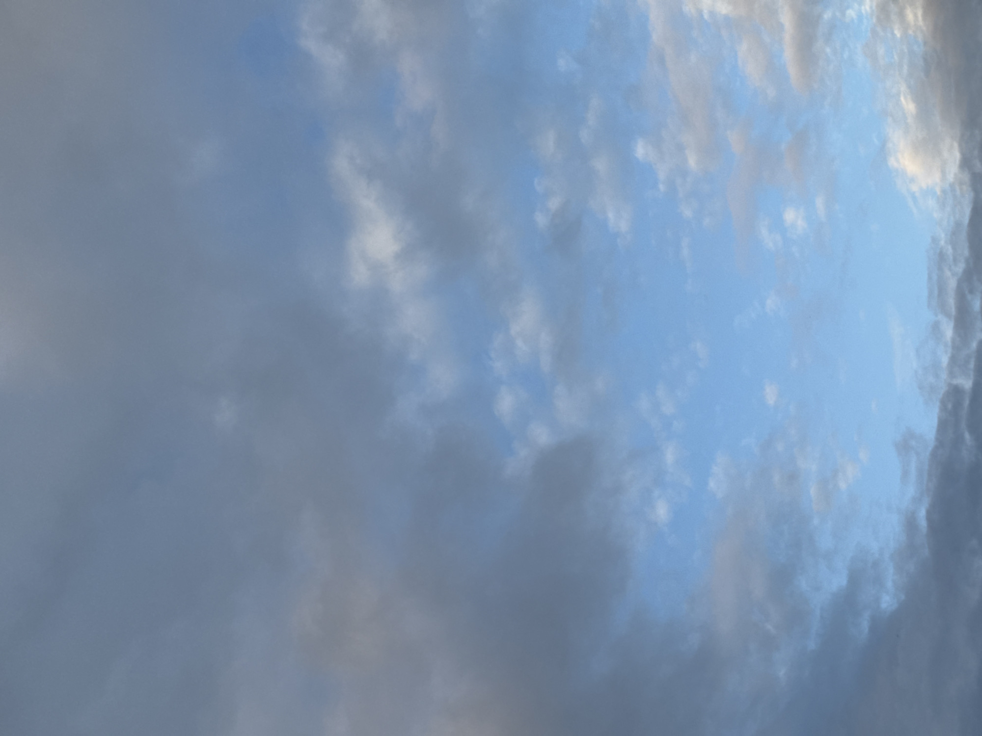 This is a vertical, full-frame photograph of a cloudy sky. The clouds are a mix of dark gray and lighter gray, with patches of bright blue sky visible between them. The clouds appear dense and heavy in some areas, while wispy and scattered in others. The overall lighting suggests it is either early morning or late afternoon. There are no discernible landmarks or signs of human activity in the image, and therefore no identifiable location within Tolyatti, Russia. The image is an abstract representation of the sky.