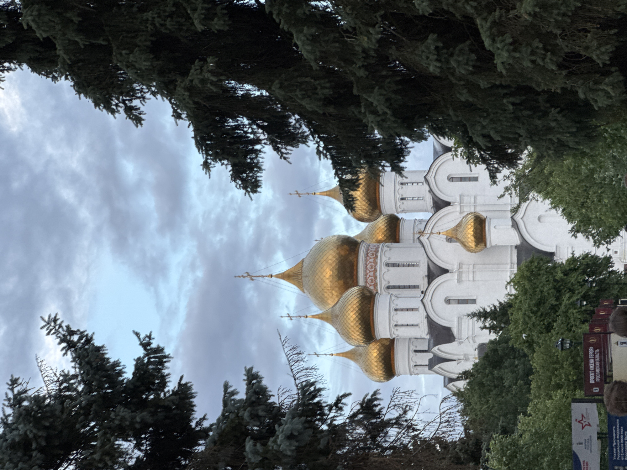 This media file shows a view of a white building with golden domes, characteristic of Russian Orthodox churches, likely in Шебунино, Russia. The building features multiple onion domes topped with crosses, and its architecture includes arched windows and decorative friezes. The structure is partially obscured by dark green coniferous trees framing the foreground and midground. The sky above is overcast with grayish clouds, suggesting a cloudy day.
In the lower right portion of the image, there are signs with Russian text. One sign reads "ПРОЕКТ «ЖЕНА ГЕРОЯ» • ЯРОСЛАВСКАЯ ОБЛАСТЬ," which translates to "PROJECT 'HERO'S WIFE' • YAROSLAVL REGION." Below this, another sign displays the Russian Federation's presidential emblem and text that appears to refer to a decree by the President of the Russian Federation. There are also glimpses of people, their heads and shoulders visible at the very edge of the frame, suggesting they are observing the scene or passing by. The overall impression is one of historical and cultural significance, set against a backdrop of natural foliage under a subdued sky. The orientation of the image is such that the golden domes are positioned vertically.