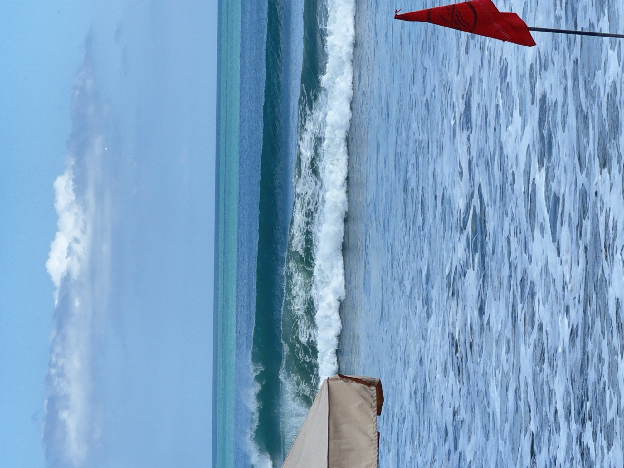 The image depicts a serene ocean scene with gentle waves rolling towards the shore. In the foreground, a portion of a beige beach umbrella is visible on the left, providing shade. To the right, a red flag on a pole stands planted in the water. The ocean is a vibrant turquoise, transitioning to a deeper blue as it extends towards the horizon. Wispy clouds dot the clear blue sky. This image was captured in Kuta, Bali, Indonesia, a popular destination known for its beautiful beaches and surfing spots. The presence of the flag might indicate a warning or a designated area, typical of beach safety measures in such locations.