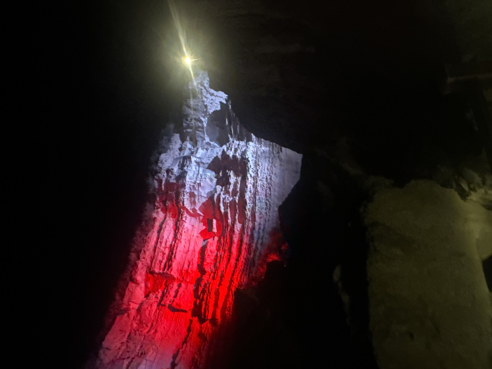 This image shows a view inside a cave, possibly a natural formation or a man-made structure adapted for exploration. The cave walls are illuminated with colored lights, predominantly red on the left side and a mix of white and bluish light on the right. The rock formations appear layered and rugged, with visible textures and crevices. A bright, focused light source, potentially a spotlight or a flashlight, is visible on the upper right side of the image, casting a strong glare. The foreground and lower parts of the image are dark, with some dimly lit surfaces that suggest a path or floor. The overall scene suggests a tourist attraction or an exploration site within a cave environment. The location in the city of Aktau, Kazakhstan, is not visually represented in the image itself.