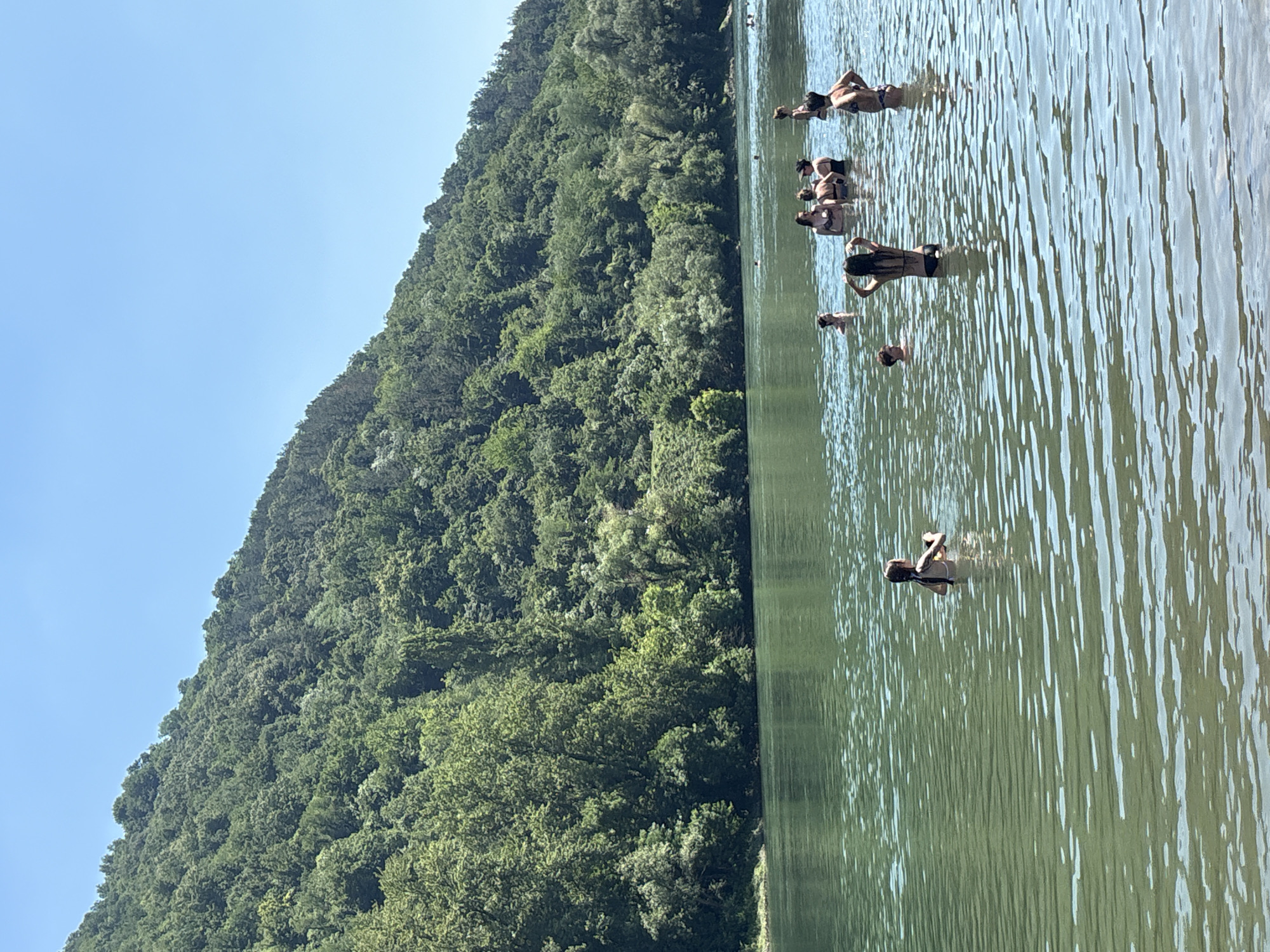 The image depicts a group of people swimming and enjoying themselves in a body of water, likely a river or lake. The scene is set against a backdrop of a densely forested, steep hillside. The water is calm with ripples reflecting the sunlight, suggesting a clear and bright day. The sky is a vivid blue, with no clouds visible, indicating pleasant weather. The time of day appears to be midday or early afternoon, given the strong sunlight.
Several individuals are visible in the water, some are standing and others are partially submerged. Their clothing appears to be swimwear, suggesting they are engaged in recreational swimming. The people are dispersed throughout the water, with no particular grouping or activity beyond general enjoyment of the water. The lush greenery of the hillside suggests a natural, possibly rural or semi-rural, environment. The presence of a forested hillside and a large body of water in Zvornik, Bosnia and Herzegovina, points to a location that is likely popular for outdoor recreation during warmer months. No specific objects other than the people themselves are prominent. There is no visible text in the image. The overall mood conveyed is one of relaxation and summer enjoyment.