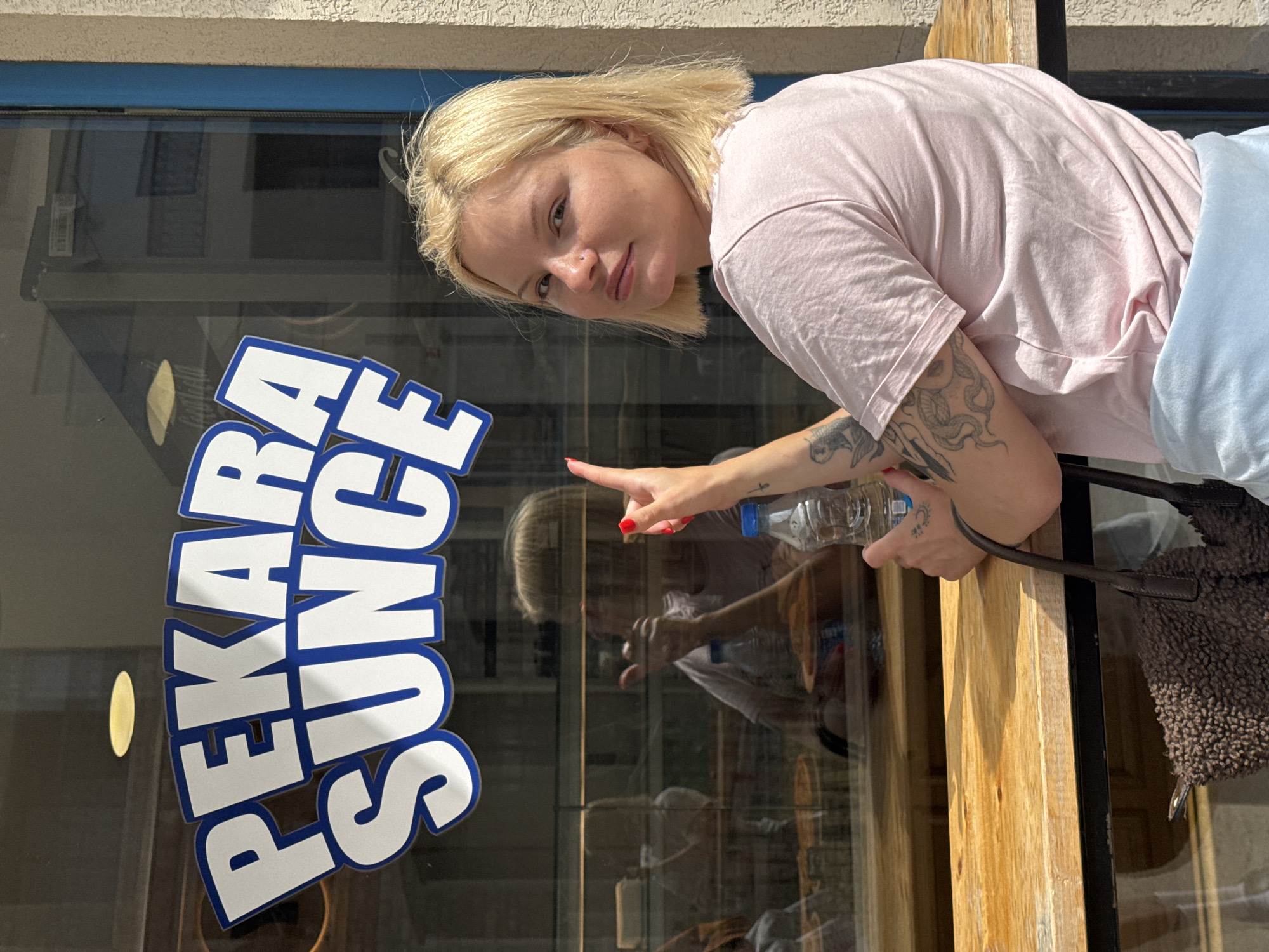 A light-skinned woman with short, blonde hair, a nose piercing, and tattoos on her arm, including a snake design, is positioned to the right of a glass window. She wears a light pink t-shirt and light blue pants. She points at the sign "PEKARA SUNCE", in blue and white letters on the window. She holds a water bottle. She has a dark-colored, fluffy purse over her shoulder. Her reflection is visible in the window. The location is the city of Petrovac centar, Montenegro.