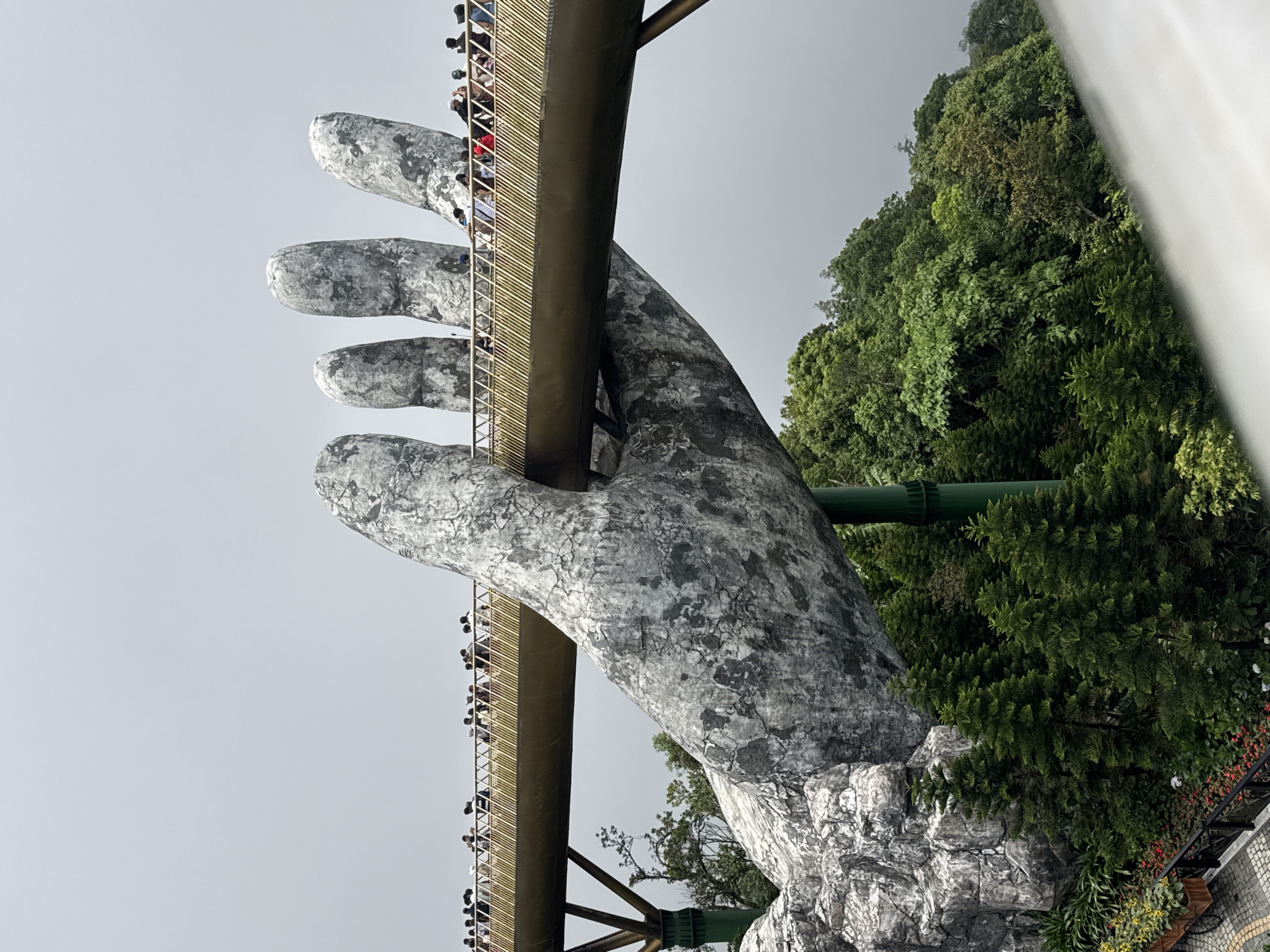 The image shows the Golden Bridge in Đà Nẵng, Vietnam. A large stone hand supports the pedestrian bridge. People are walking along the bridge. Lush green vegetation surrounds the structure. The sky is overcast.