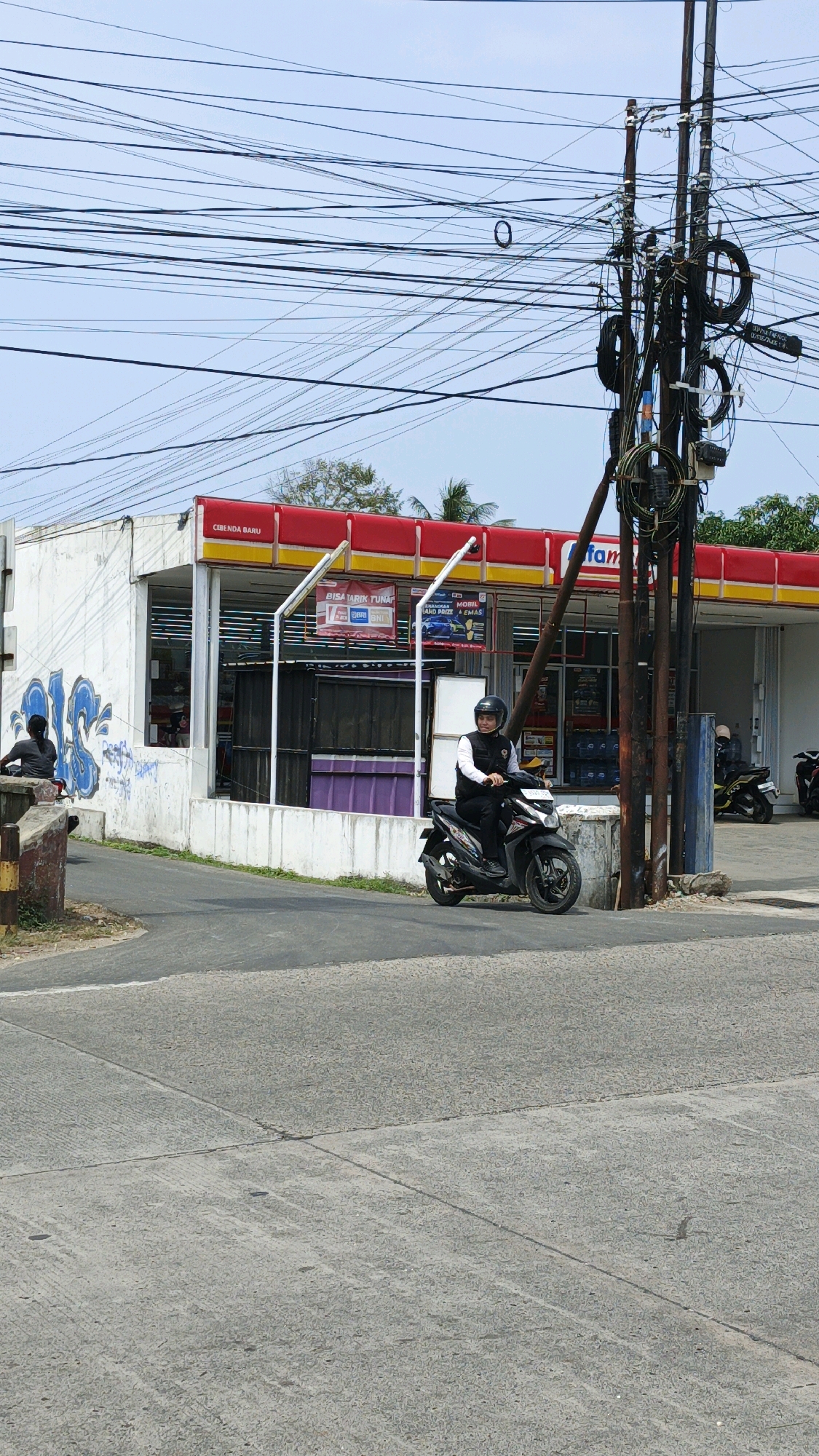 A person wearing a black helmet and a white vest over a black shirt rides a black motorcycle on a paved road in front of a minimarket in Cibenda, Indonesia. The minimarket has a red and yellow awning and a white facade with graffiti on the left side. Several utility poles with a dense network of wires are visible behind the minimarket. The sky is clear and blue, and trees are visible in the background.