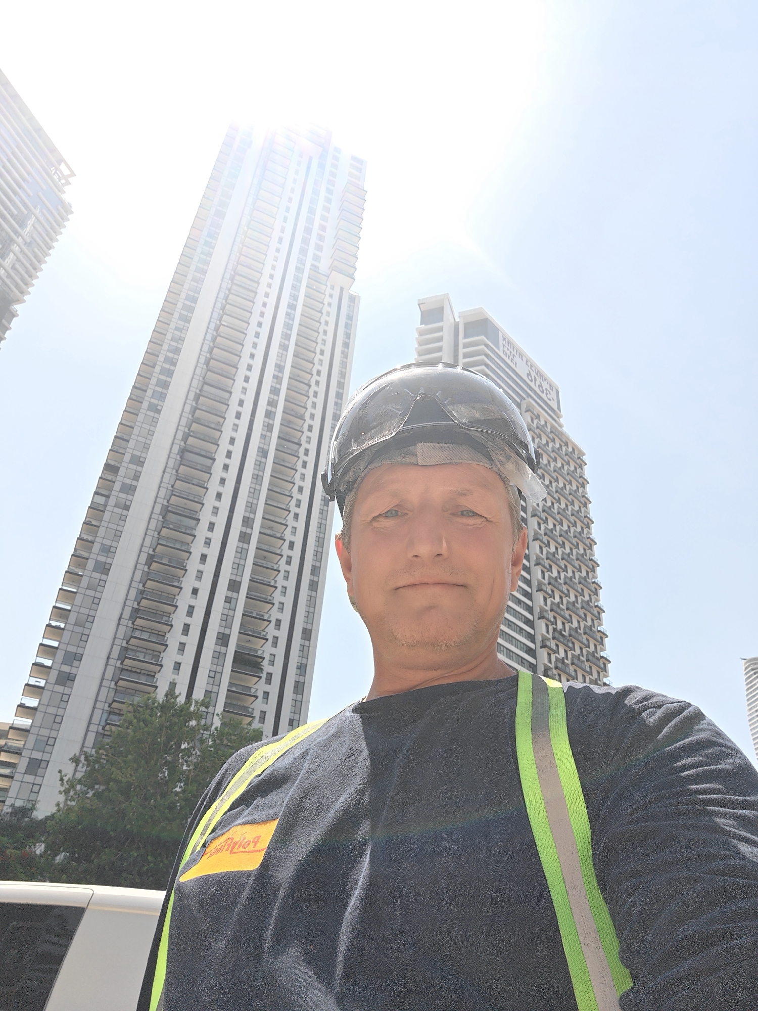 A man wearing a dark blue long-sleeved shirt, a reflective yellow and green vest, and a black construction helmet is standing in front of a tall, modern building in Givatayim, Israel. The man is looking directly at the camera, and his face is partially illuminated by sunlight. The building behind him is white with many windows and balconies, and the sky above is clear and bright. The sun is shining brightly, creating a lens flare in the upper left portion of the image.
