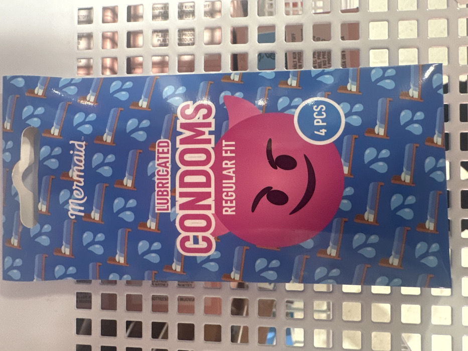 A blue, rectangular retail package for "Mermaid" brand lubricated condoms is centered in the frame, positioned in front of a white, square-grid display structure. The package features a small hanging cutout at the top. Its background design consists of a repeated pattern of small, brown-framed beds with blue bedding and white pillows, interspersed with light blue droplet shapes. White text "Mermaid" is at the top. Below this, the word "LUBRICATED" appears in white against a red horizontal bar, followed by large pink lettering spelling "CONDOMS." Beneath "CONDOMS," "REGULAR FIT" is printed in white. A large, dark pink circular character with small black eyes, a black smile, and two pointed ears (resembling a devil or imp emoji) occupies the lower central portion of the package. A smaller blue circle with the white text "4 PCS" is overlaid on the lower right of this pink character. The square grid background exhibits reflections and distorted, partially legible text fragments, suggesting the presence of other items on an adjacent shelf or within the retail environment. This scene is located in Saint-Germain-en-Laye, France.