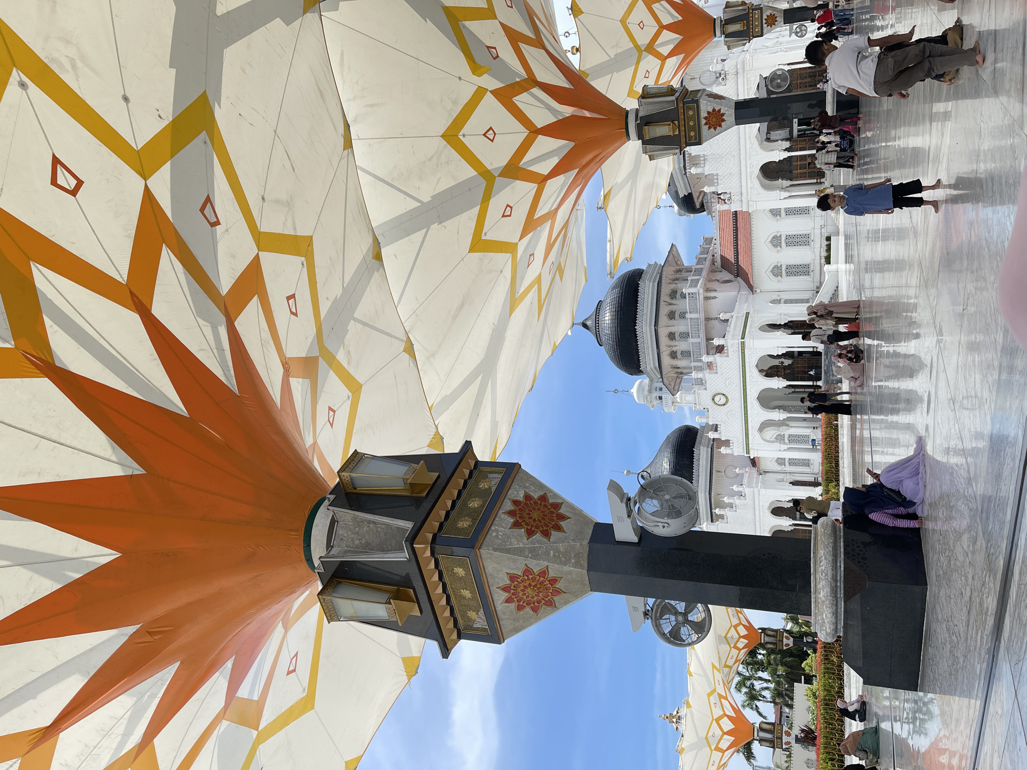 The image captures the courtyard of the Baiturrahman Grand Mosque in Banda Aceh, Indonesia, under a bright blue sky. The foreground is dominated by a large, ornately decorated umbrella structure, its white fabric canopy patterned with orange and yellow geometric designs. Beneath the canopy, a tall, dark gray pillar supports the structure and features decorative panels with red and gold floral motifs, along with two electric fans. The polished, reflective floor of the courtyard shows the reflections of the umbrellas and the mosque.
In the mid-ground, the white facade of the Baiturrahman Grand Mosque stands prominently, characterized by its multiple domes, arches, and minarets. The mosque is a significant architectural landmark. People are scattered throughout the courtyard. Some are seated on the floor, including women wearing hijabs in various colors. Others are standing and walking, appearing to be visitors or worshippers. Two young boys are visible in the center of the courtyard, one standing and looking forward, the other partially obscured and interacting with something on the ground.
The overall scene depicts a public space with architectural grandeur, cultural elements, and human activity, indicative of a place of worship and community gathering. The lighting suggests it is daytime, likely a sunny day.