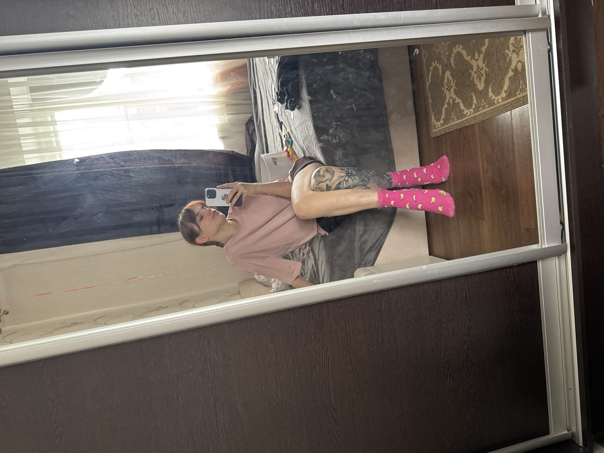 The image shows a mirror reflection of a woman lying on a bed and taking a picture of herself with her phone. She is wearing a light pink shirt and pink socks with banana patterns. Her legs are visible, and one of them has a tattoo. The bed has a grey blanket on it. In the background, there is a window with blinds and a rug on the floor. The mirror is surrounded by a frame, and the reflection shows part of a dark brown wooden surface. The image does not provide any information to identify the location as Rostov-on-Don, Russia.